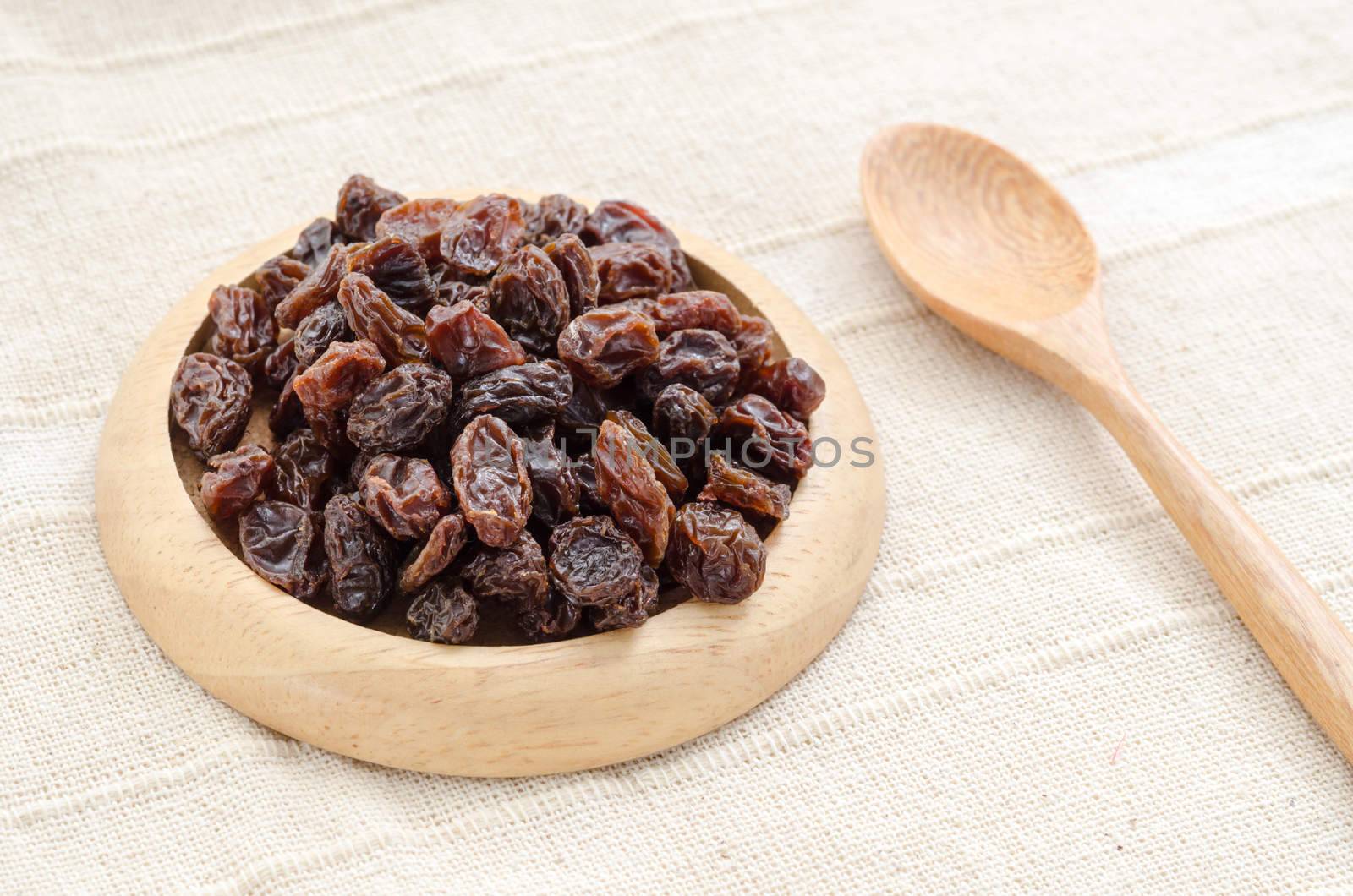Raisins in wood dish with wooden spoon on tablecloth.