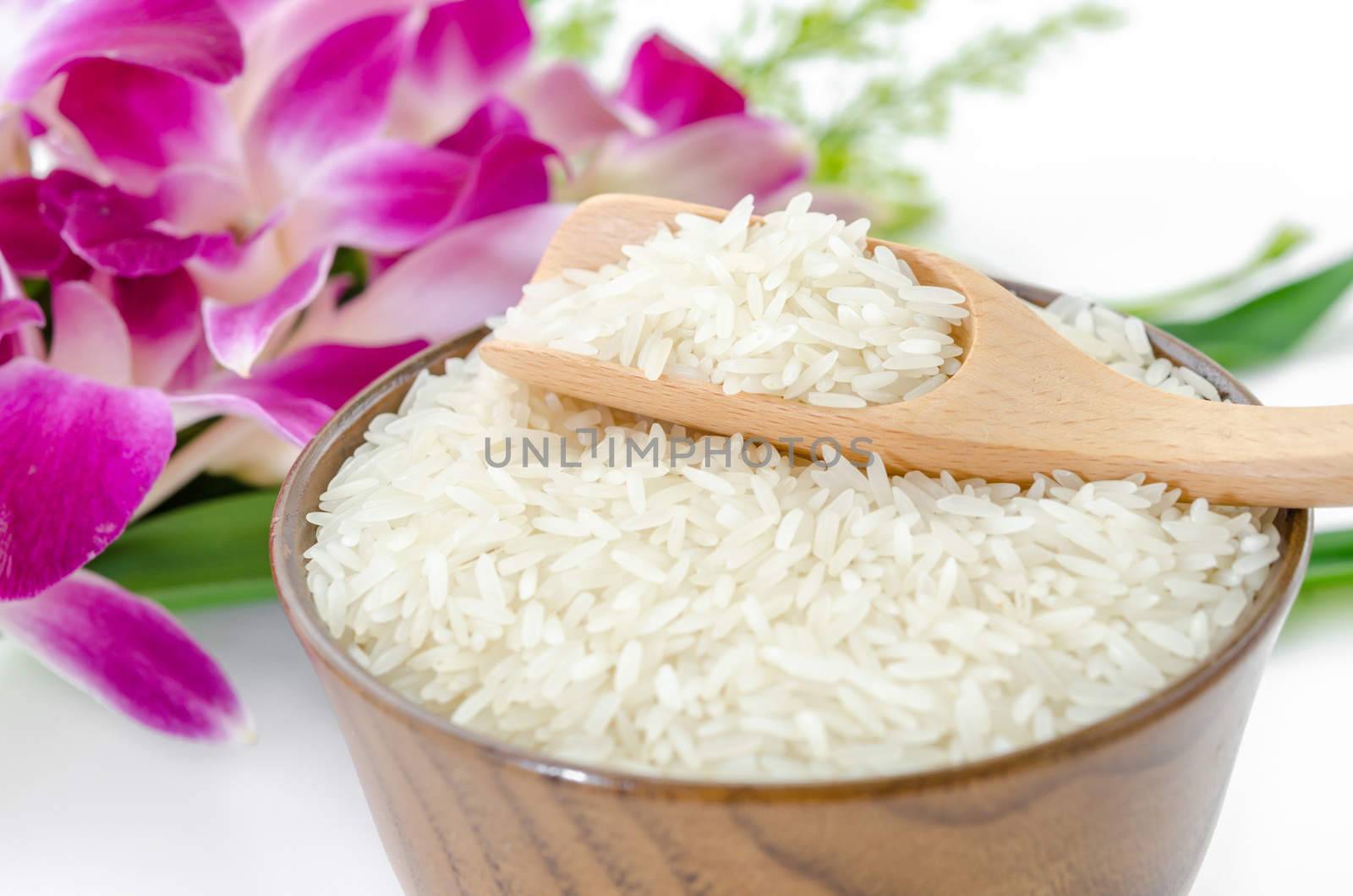 Raw rice jasmine and wooden spoon in wooden bowl with violet orchid flower on white background.