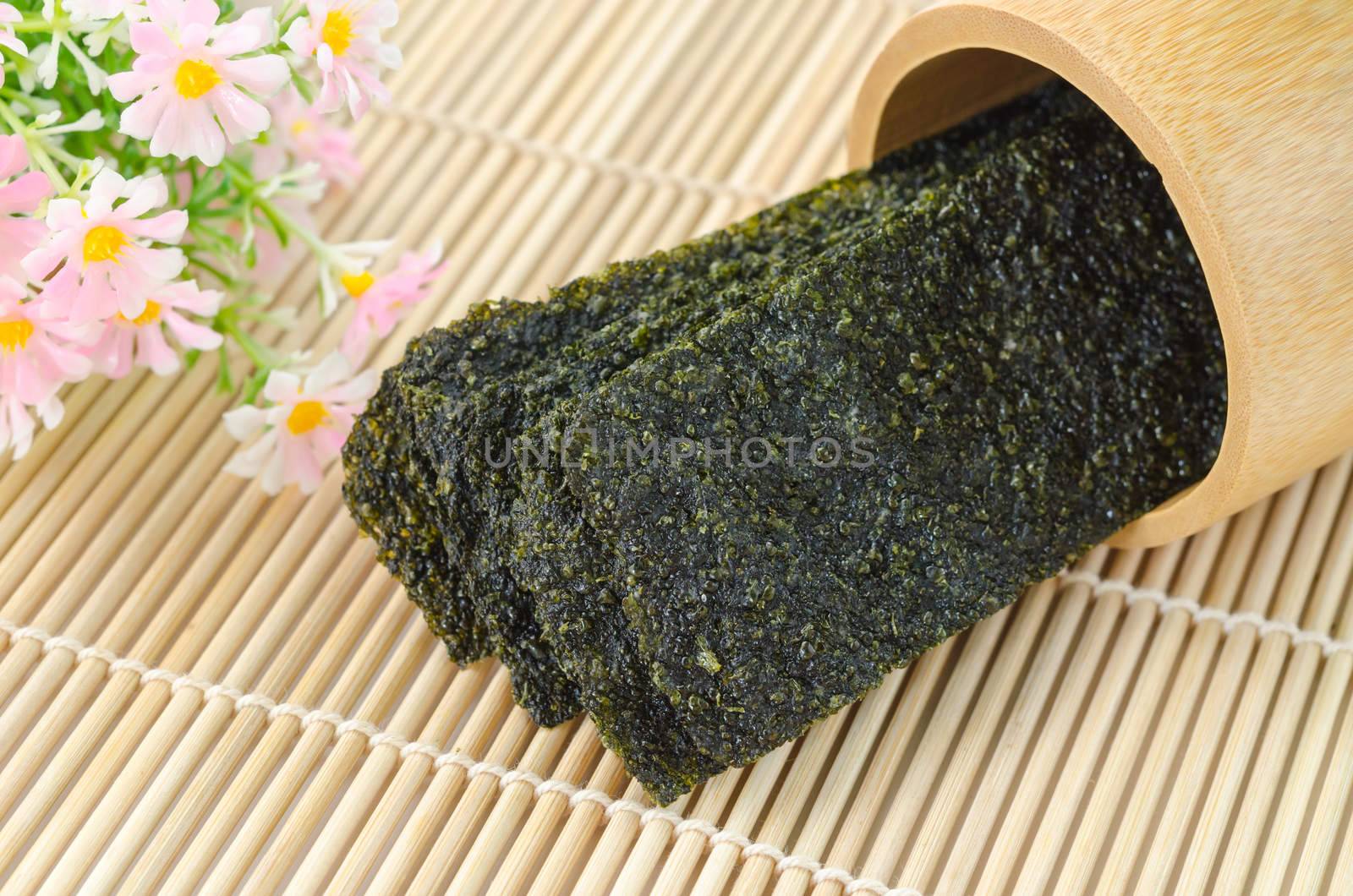 Fried seaweed in wooden cup with flower on bamboo mat.