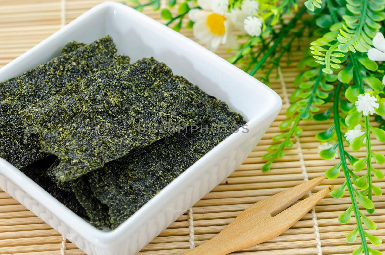 Fried seaweed in white bowl and wooden fork spoon with flower on bamboo mat.