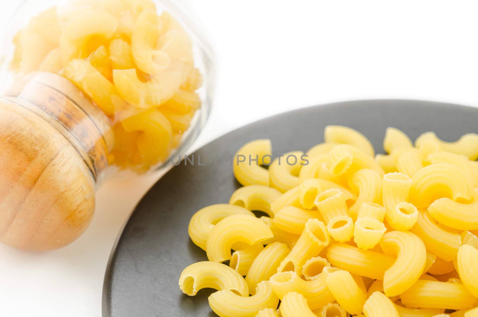 Uncooked elbow macaroni on black dish and in grass on white background.