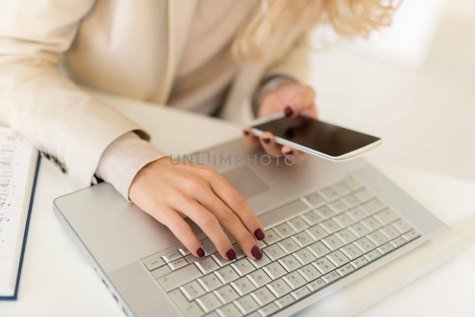 Close-up of female hand with painted nails on the keyboard of the laptop and typing on smart phone.