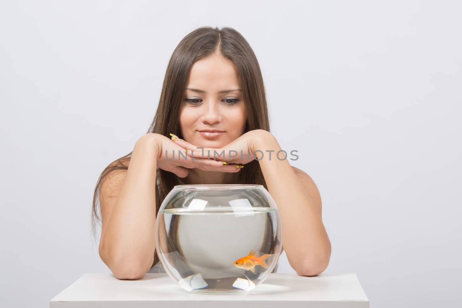 A young girl sits next to a round aquarium in which swimming goldfish