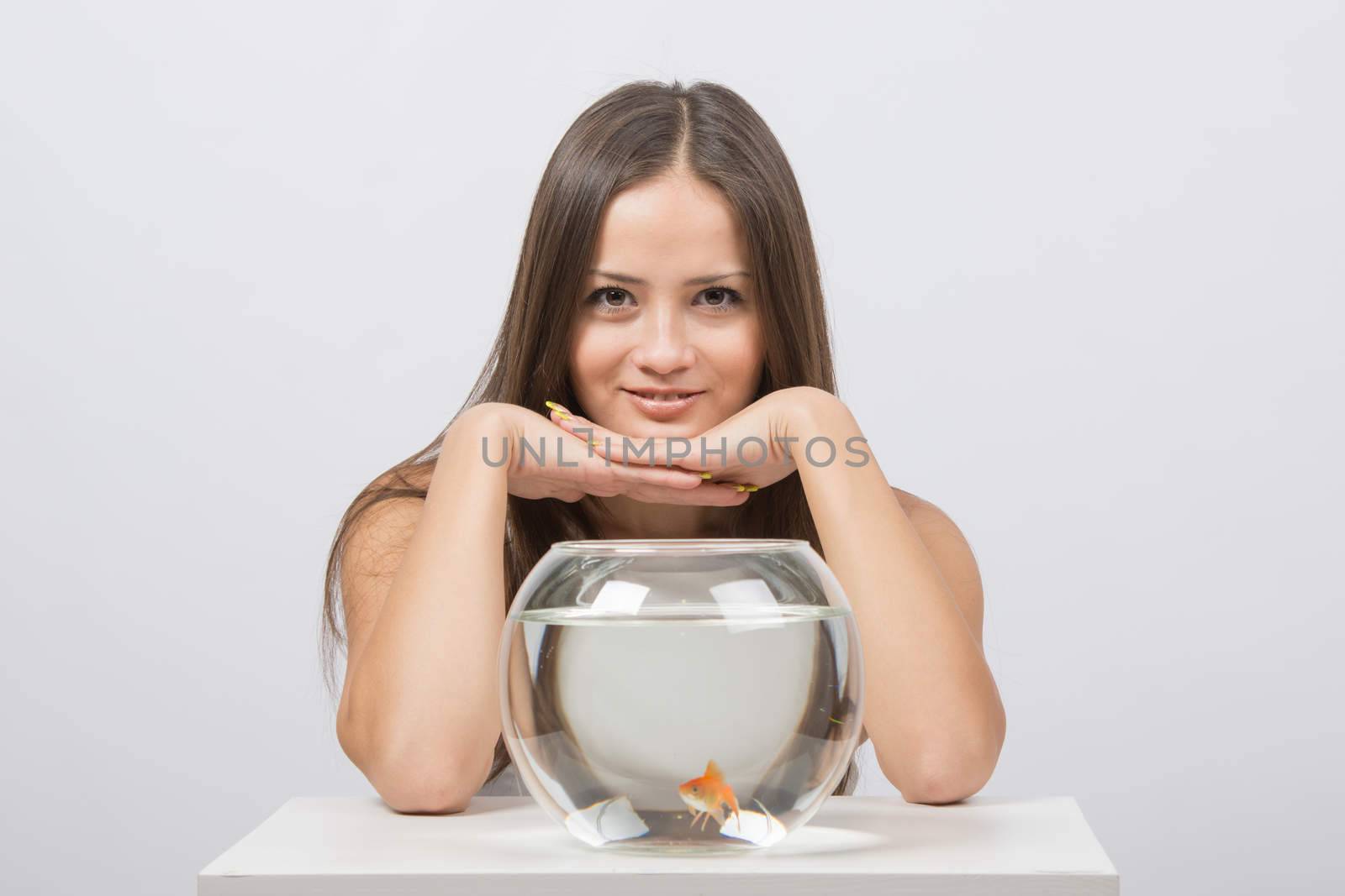 A young girl sits next to a round aquarium in which swimming goldfish