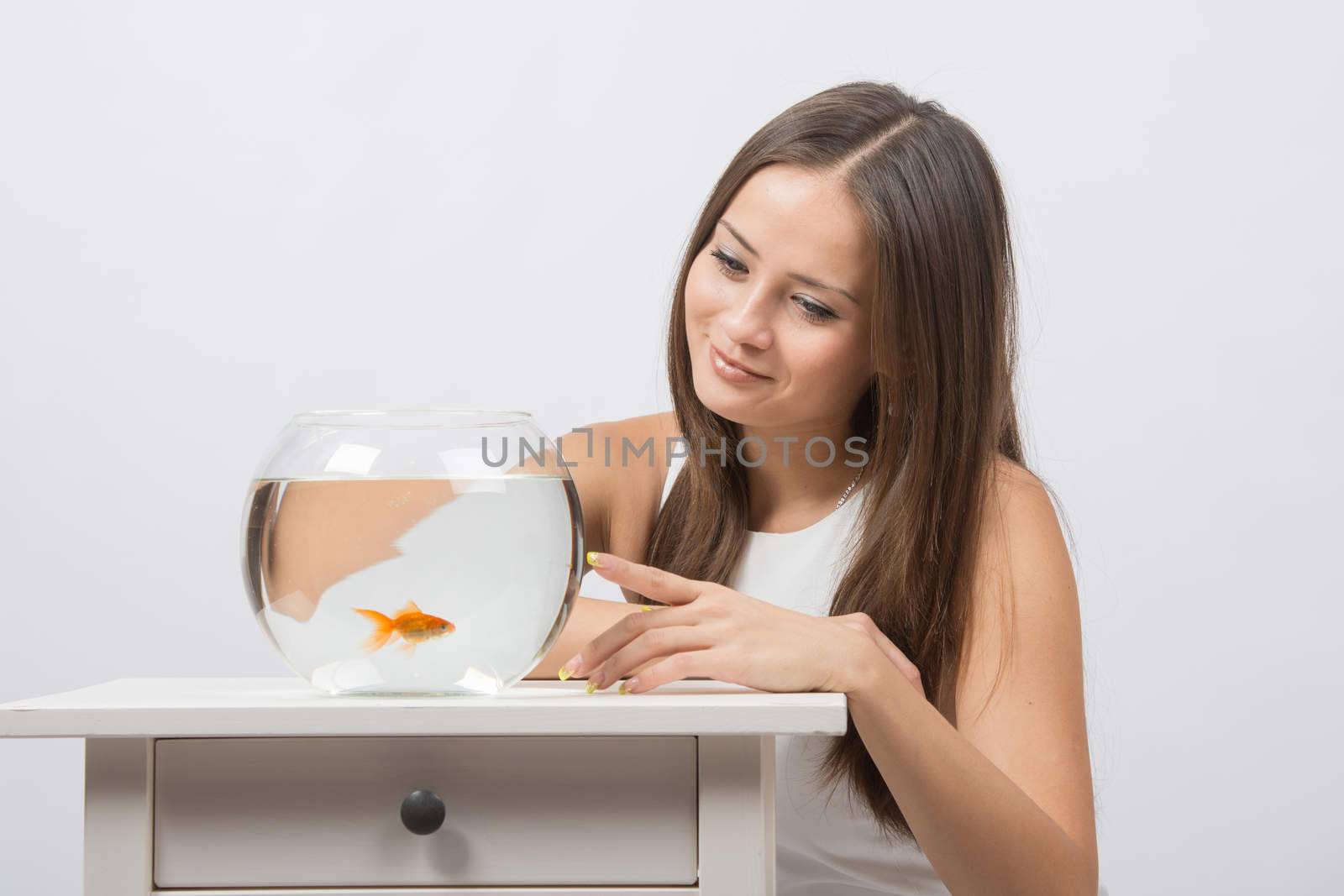 A young girl sits next to a round aquarium in which swimming goldfish