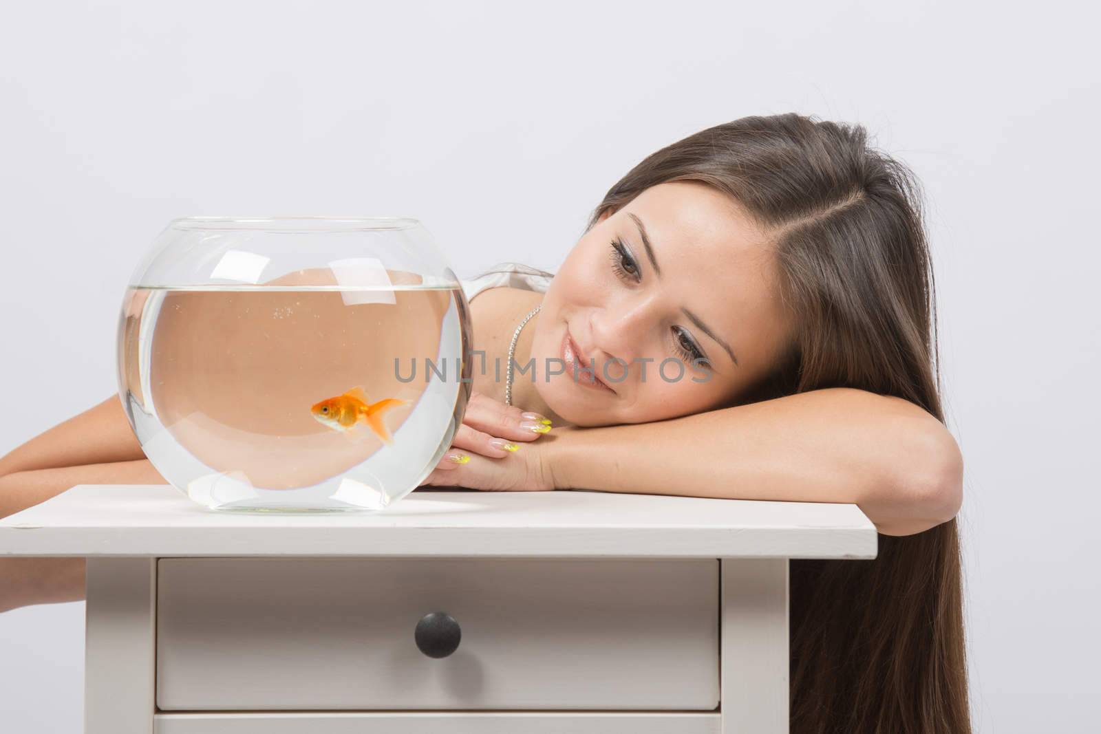 A young girl sits next to a round aquarium in which swimming goldfish