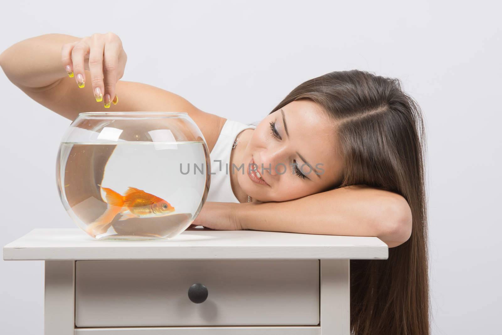 A young girl sits next to a round aquarium in which swimming goldfish