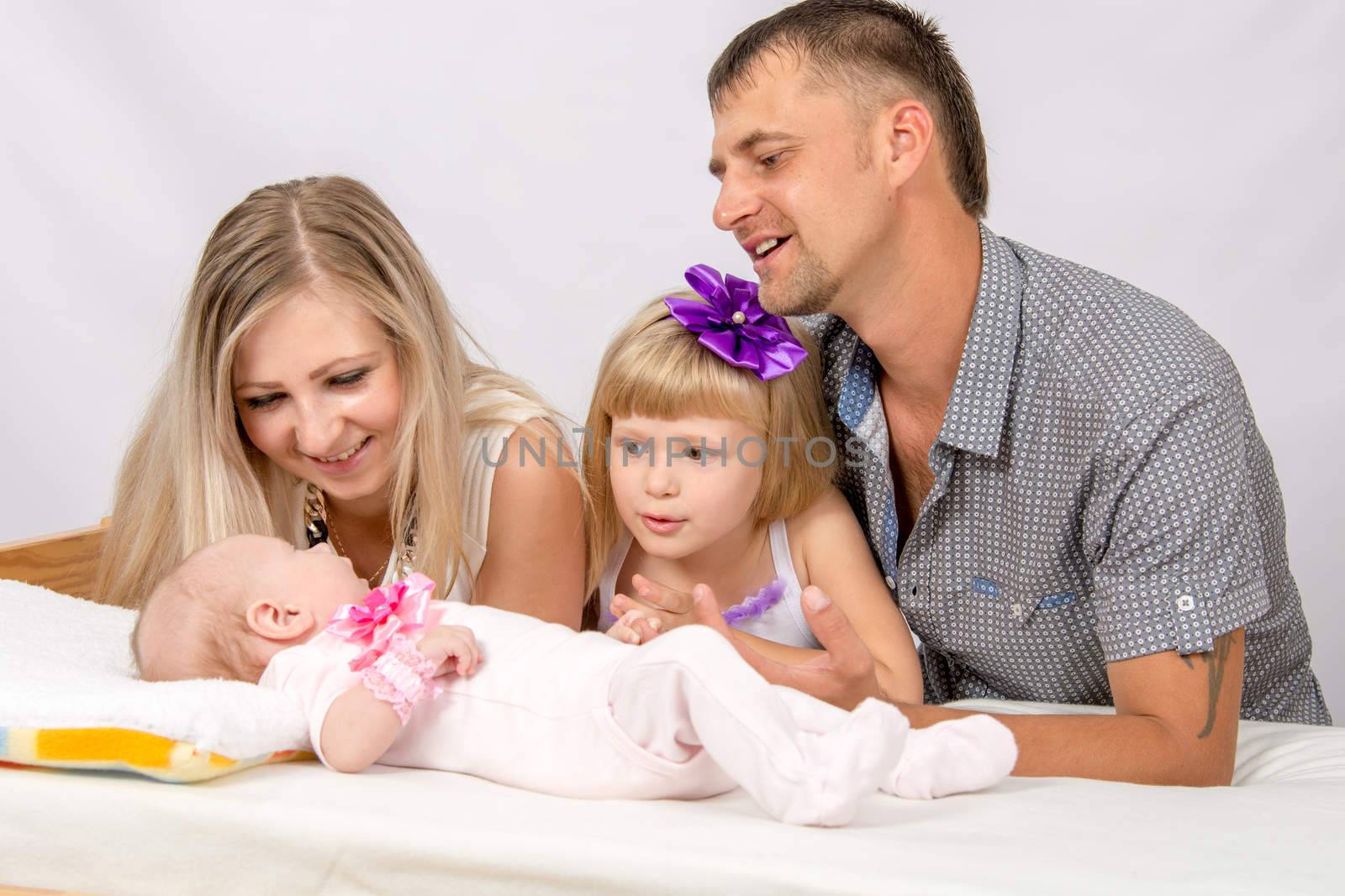 Young parents and daughter sit at the five-year crib, the crib is on a two-month baby on her back