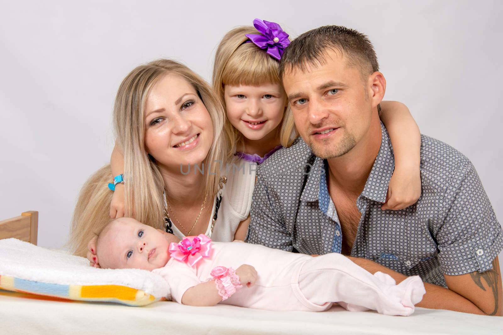 Young parents and daughter sit at the five-year crib, the crib is on a two-month baby on her back