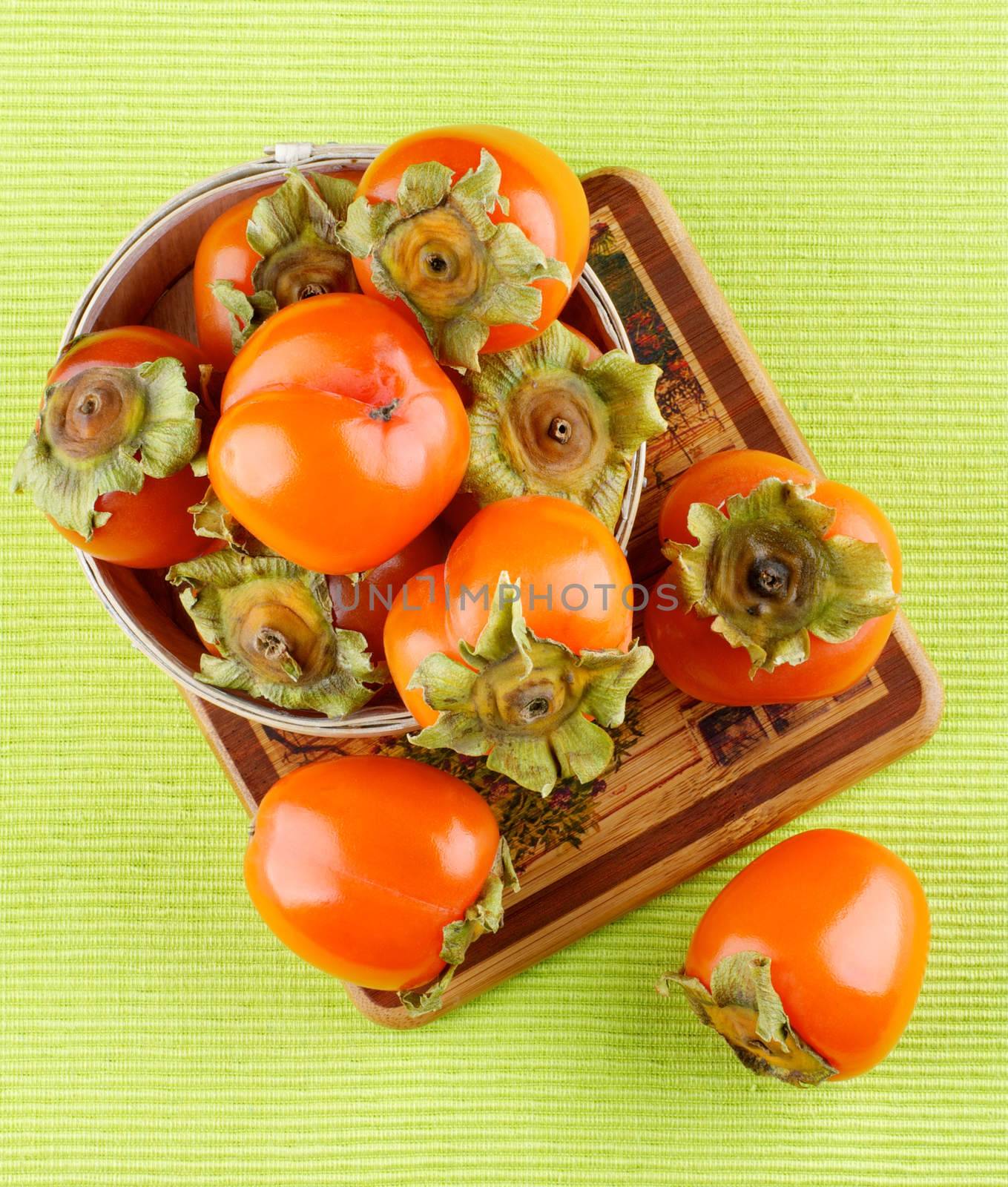 Heap of Delicious Raw Persimmon in Wooden Bowl closeup on Green Textile Napkin background. Top View