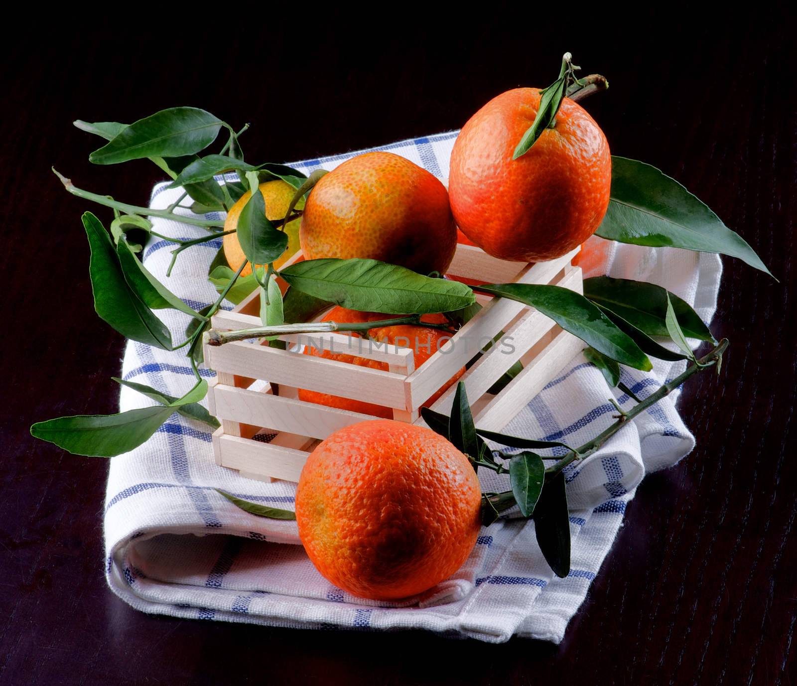 Arrangement of Ripe Tangerines with Leafs in Wooden Box on Napkin closeup on Dark Wooden background