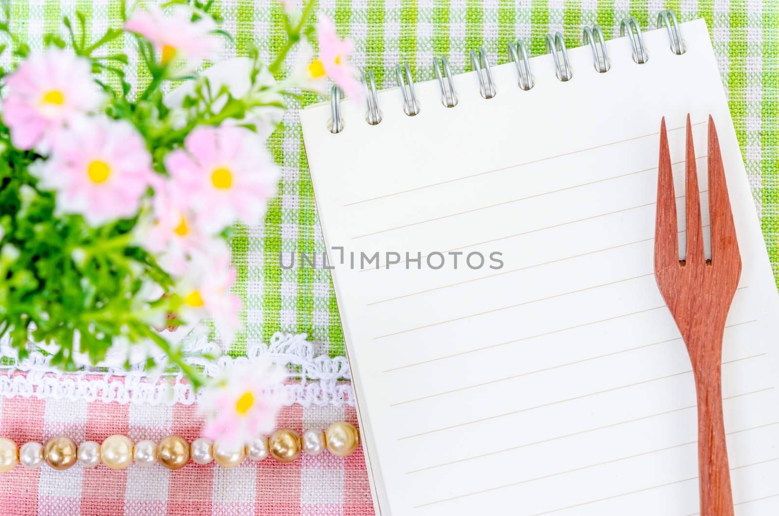 Open diary and wooden spoon fork with flower on beautiful fabric background. Menu concept.