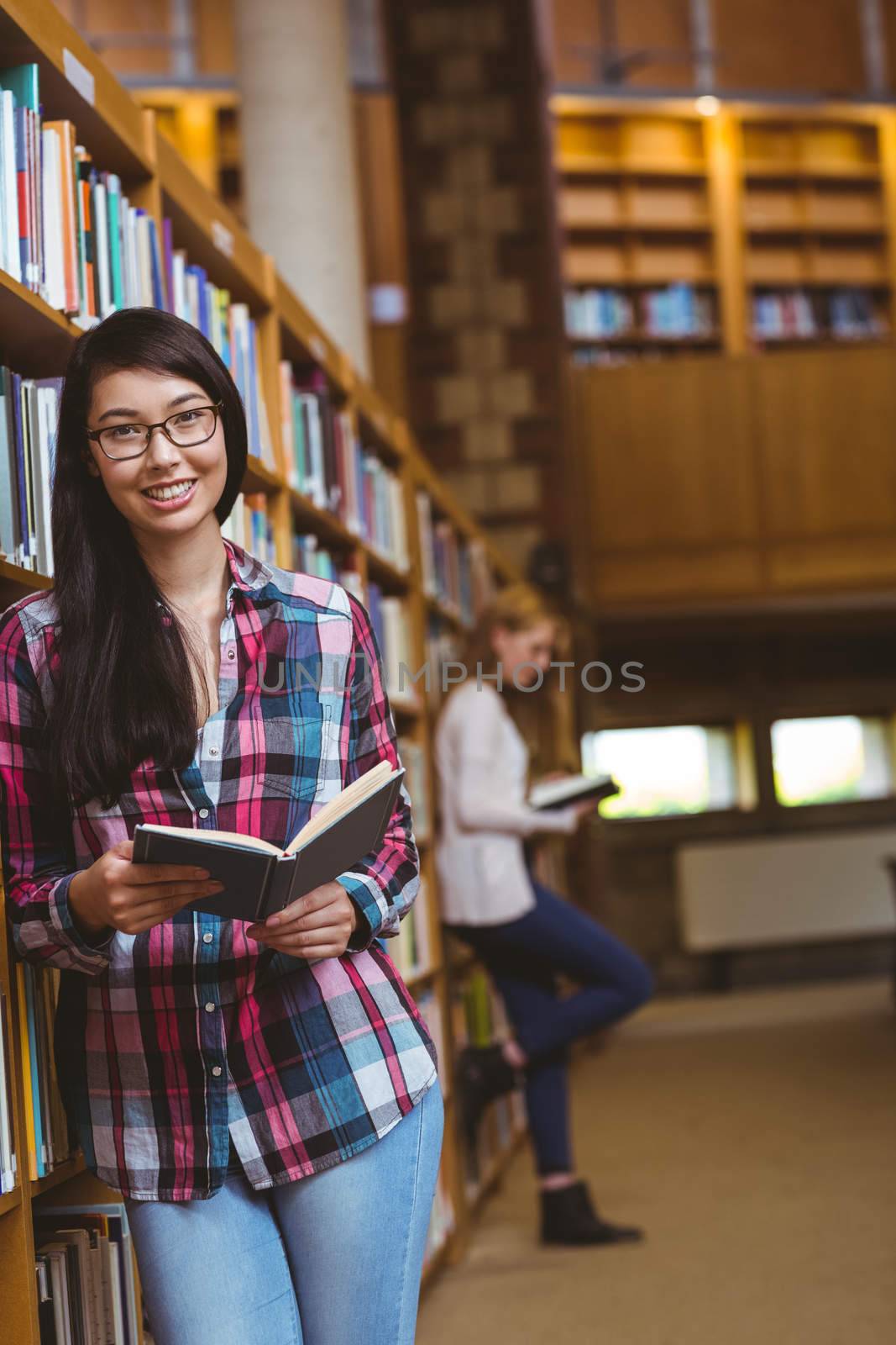 Smiling student leaning against bookshelves reading book at the university
