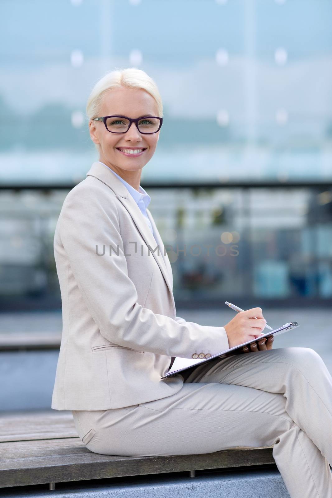 business, people and education concept - young smiling businesswoman in glasses with notepad over office building