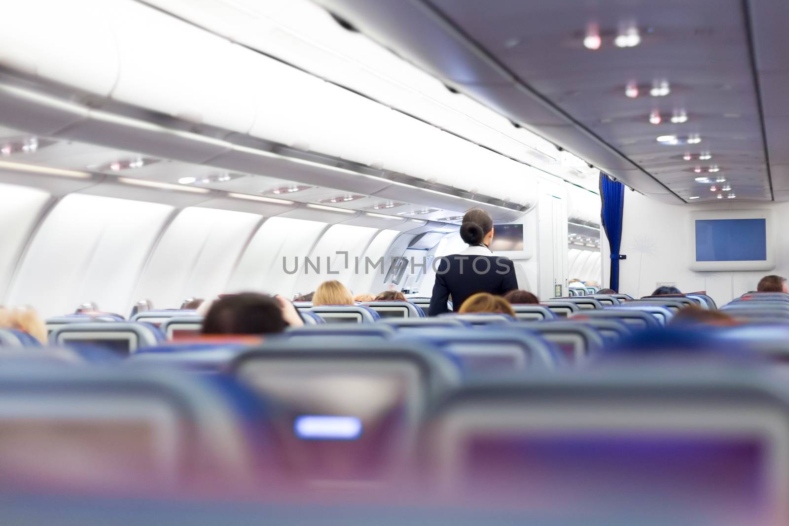 Interior of large passengers airplane with people on seats and stewardess in uniform walking the aisle. 