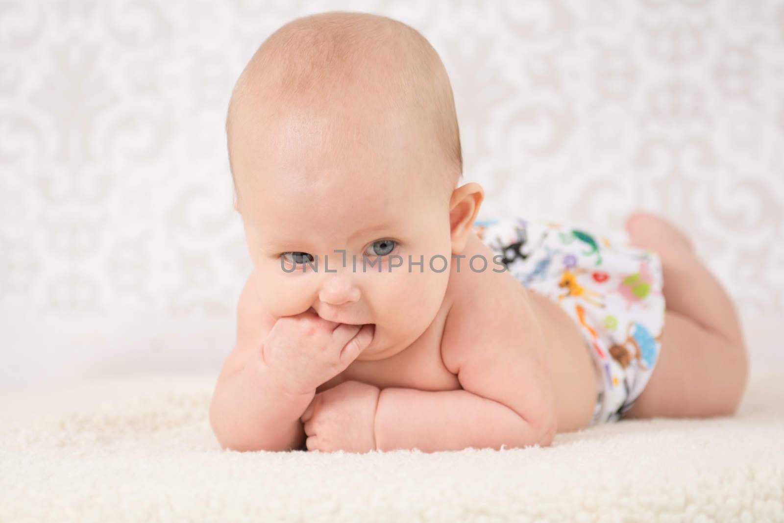 Baby in a reusable nappy, lying on a belly on a light background, bitting her fist