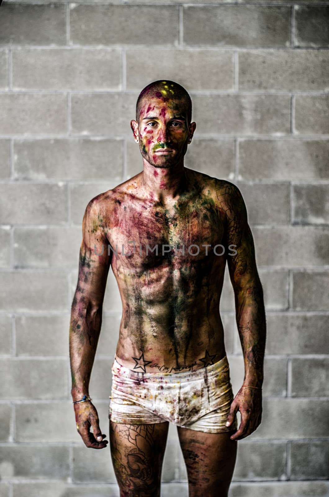 Handsome young man with skin painted all over with bright Holi colors, in front of concrete bricks wall