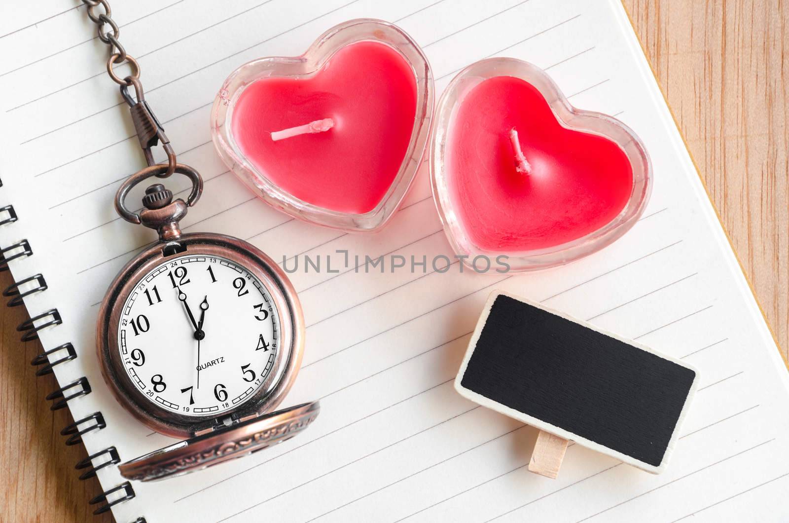 Red heart candle and pocket watch with open diary on wooden background.