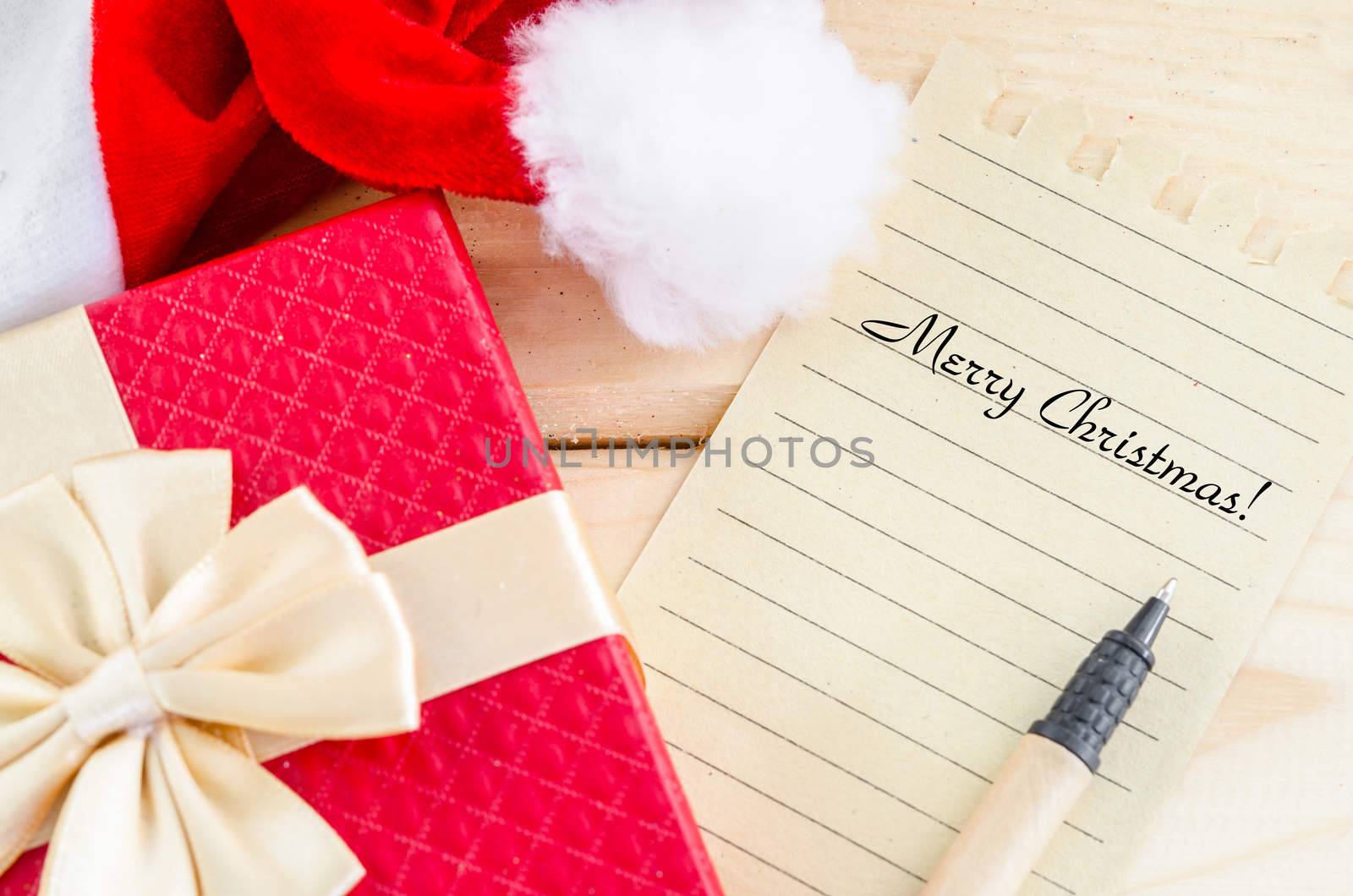 Merry Christmas word writing with pen on brown paper with red gift box on wooden background.