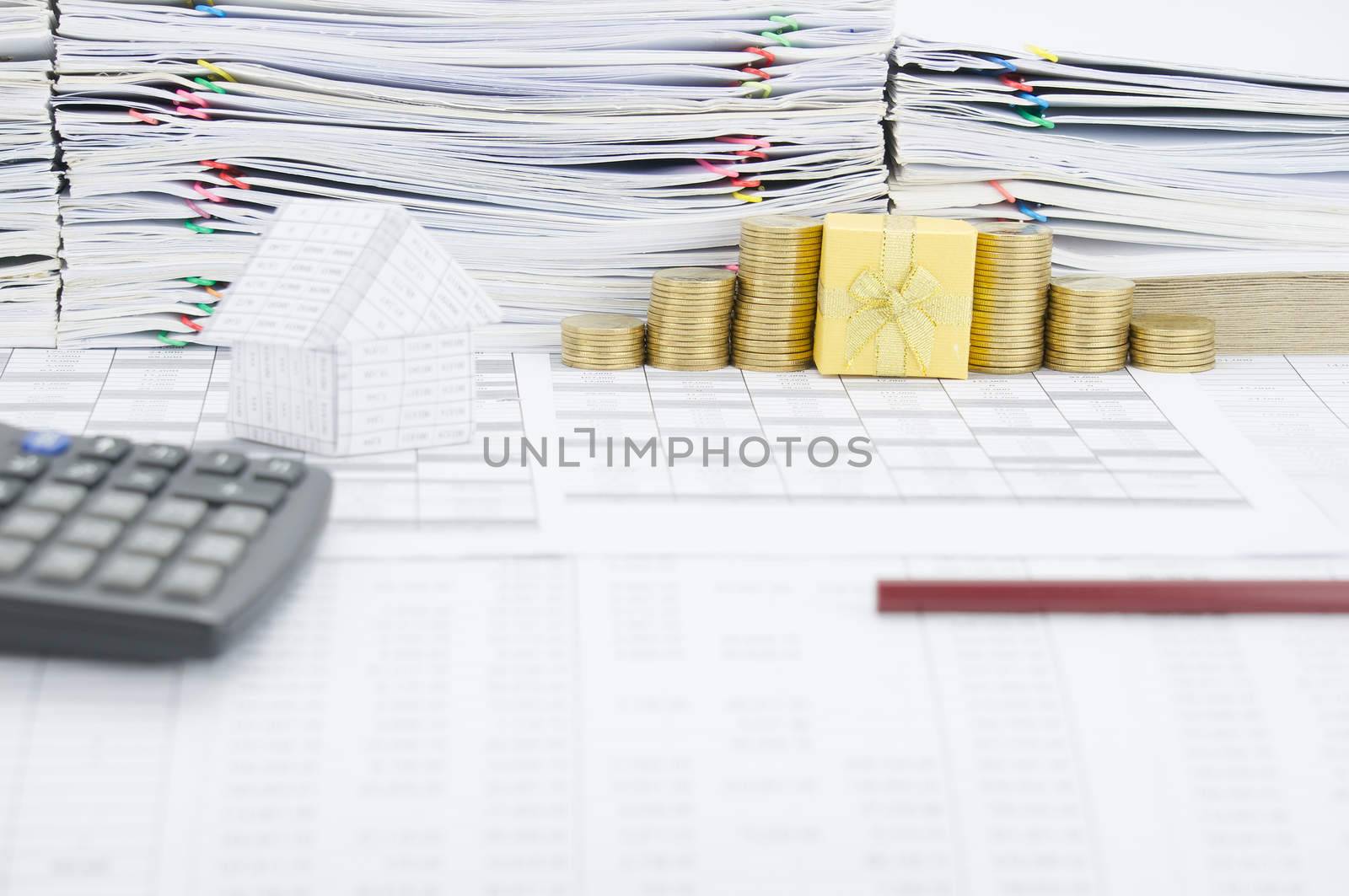 Gold gift box between pile of gold coins on finance account have blur house with calculator and brown pencil as foreground and pile of paperwork as background.