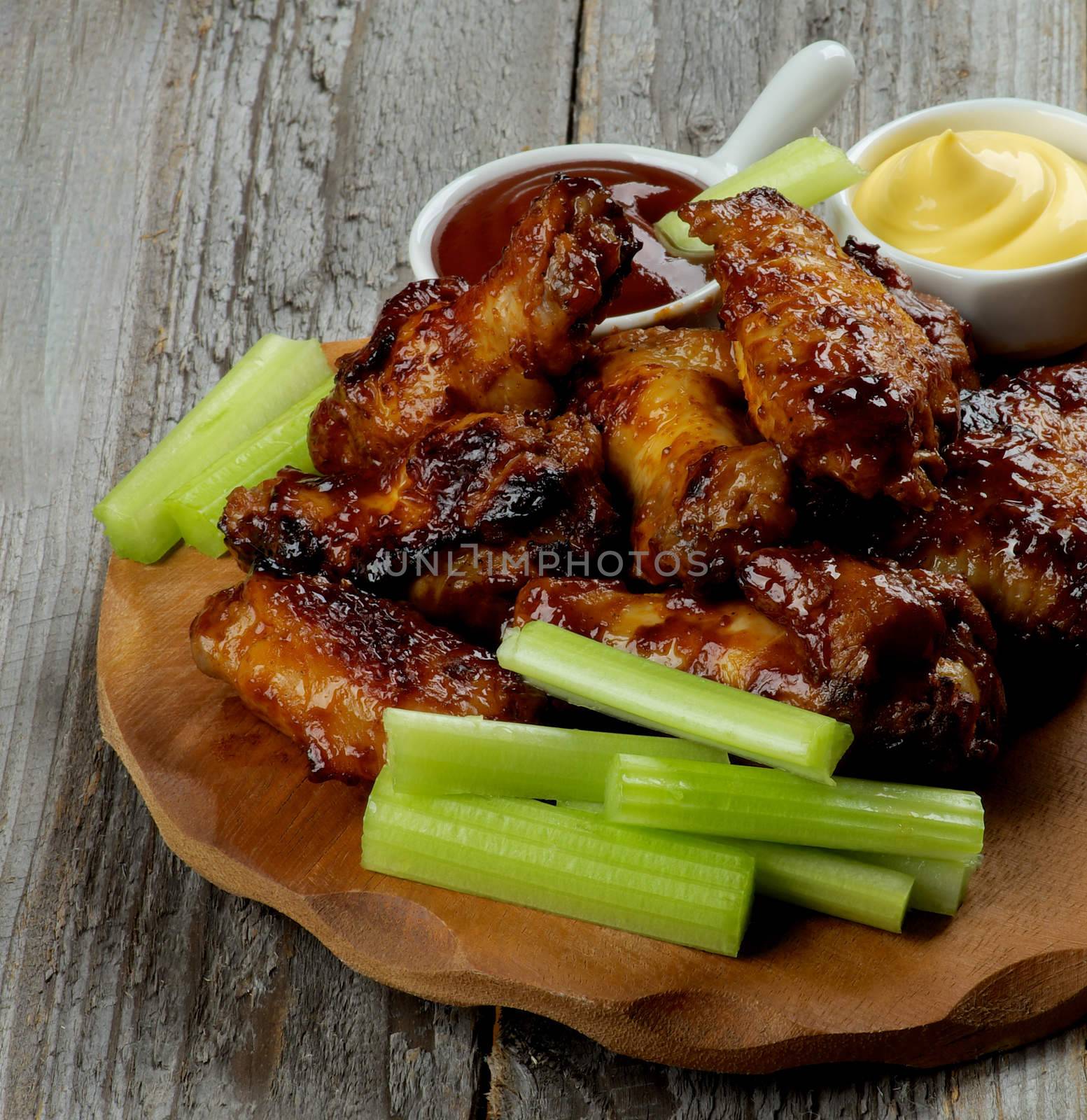 Delicious Chicken Legs and Wings Barbecue with Ketchup and Cheese Sauces and Celery Sticks on Wooden Plate closeup on Rustic Wooden background