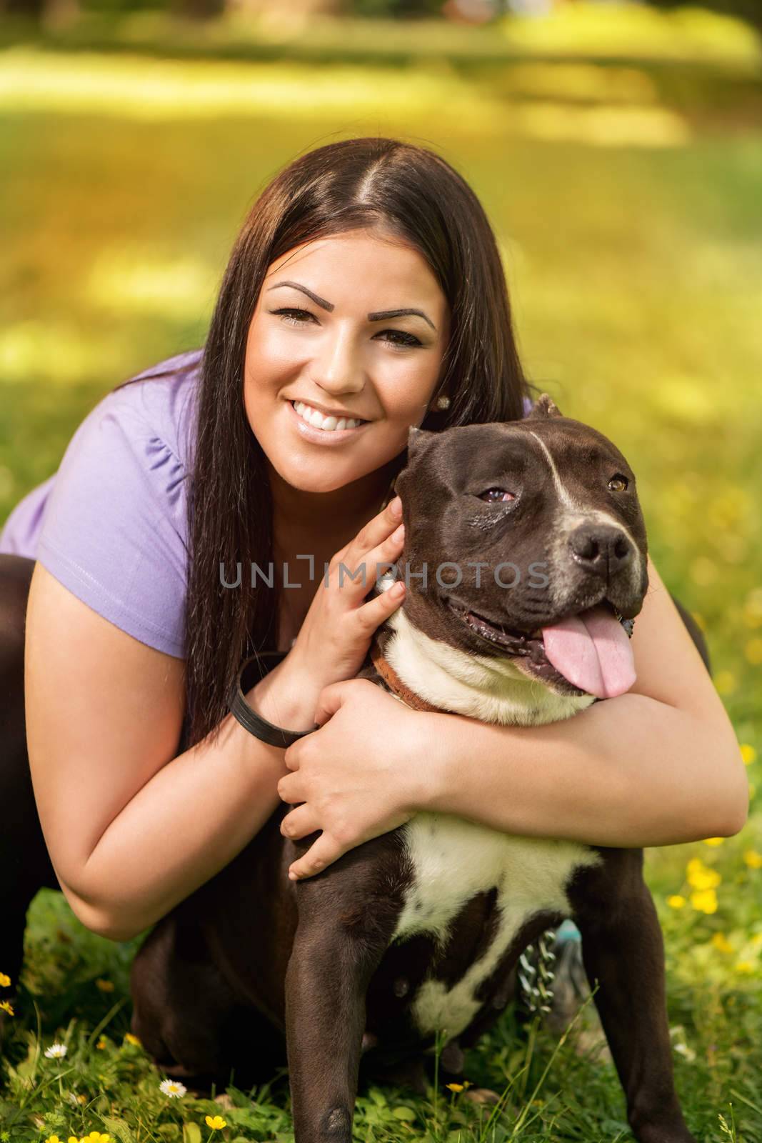 Portrait of a beautiful young woman hugging her cute stafford terrier. They sitting on grass in the park and enjoying. Looking at cemera.
