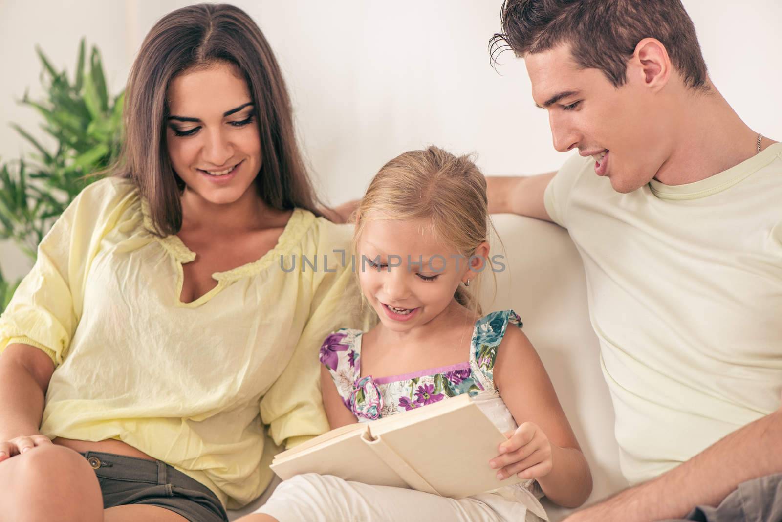 Beautiful Happy young mother and father and their cute daughter sitting at home and reading book