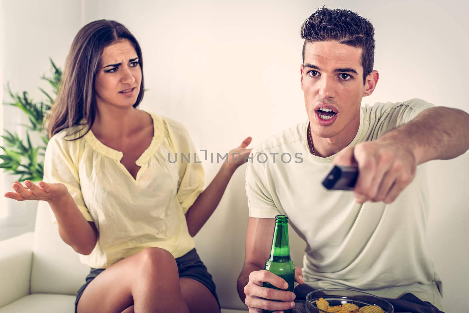 Young man sitting on sofa and watching tv. His angry wife yelling.