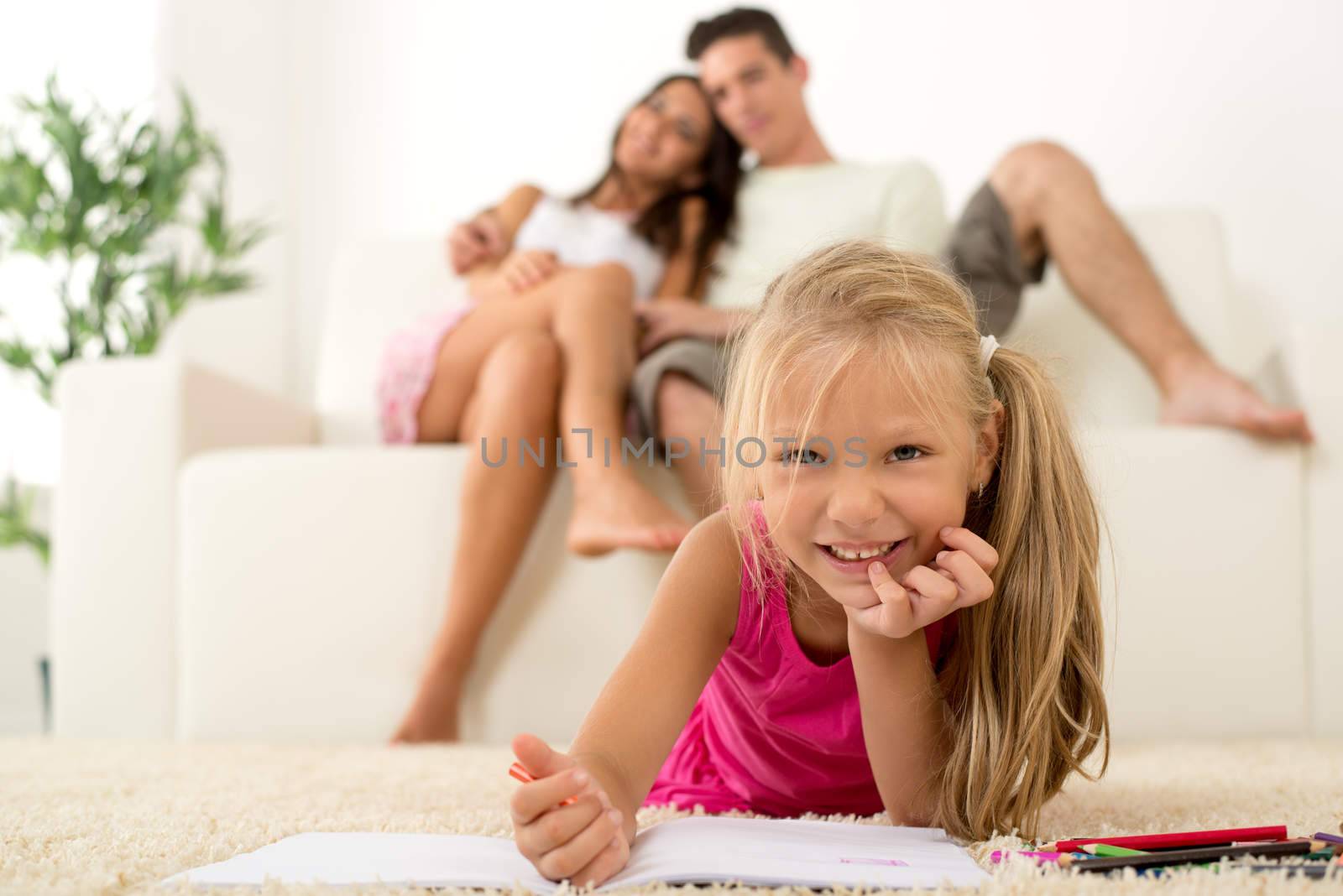 Happy family at home. Mother and father sitting, their cute little girls lying down and drawing with colored pencils. Focus On little girl.