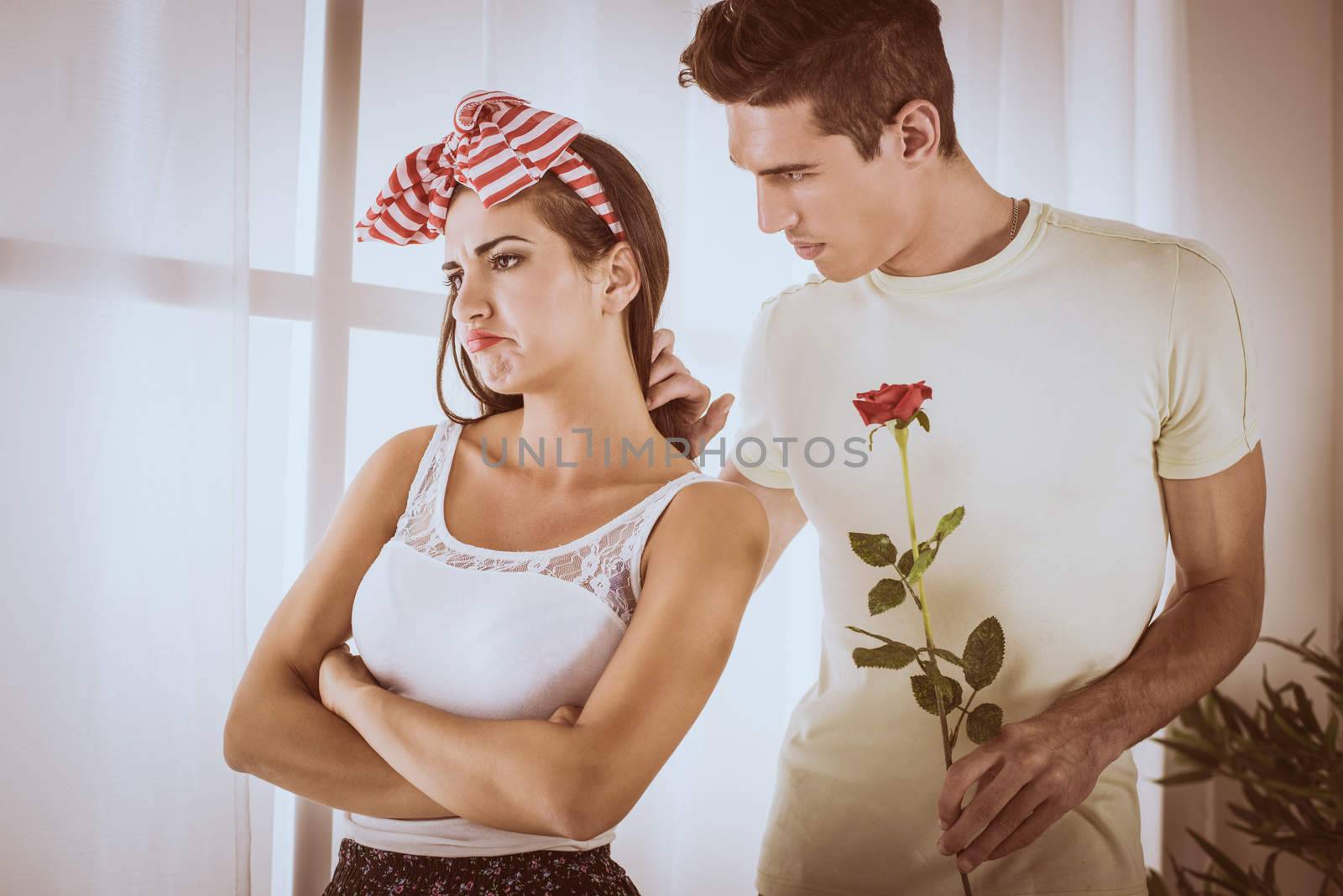 Displeased beautiful girl standing not interested while he is cute boyfriend in love holding rose.