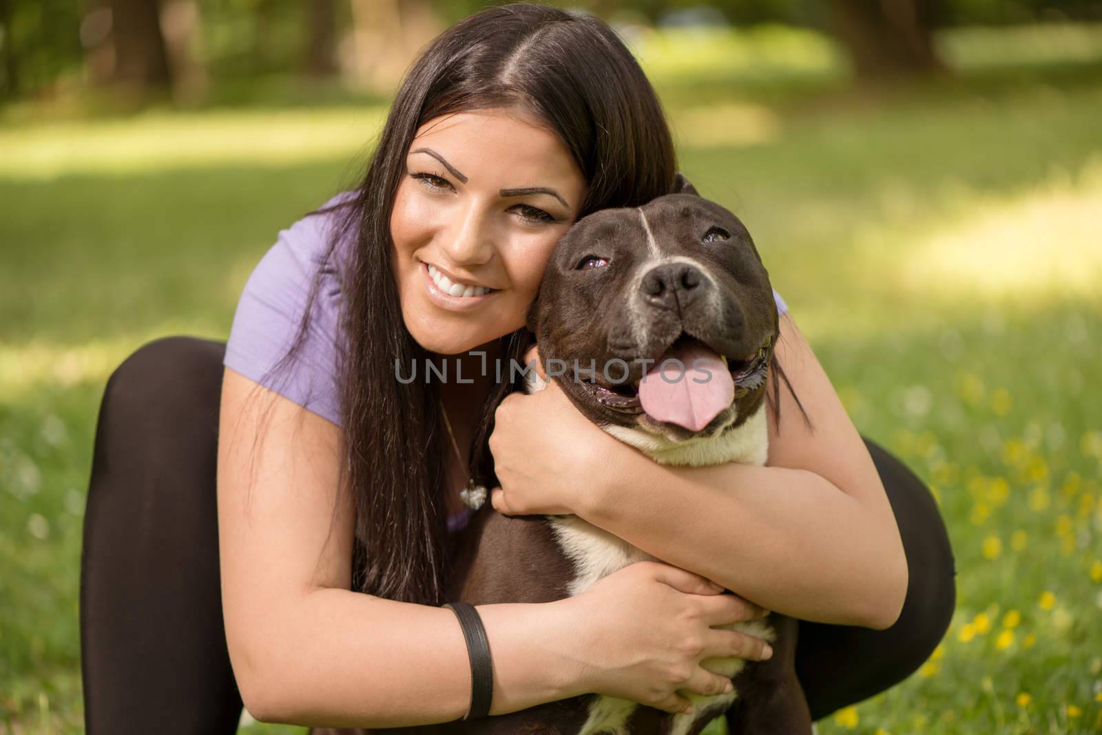 Portrait of a beautiful young woman hugging her cute stafford terrier in the park and enjoying. Looking at cemera.