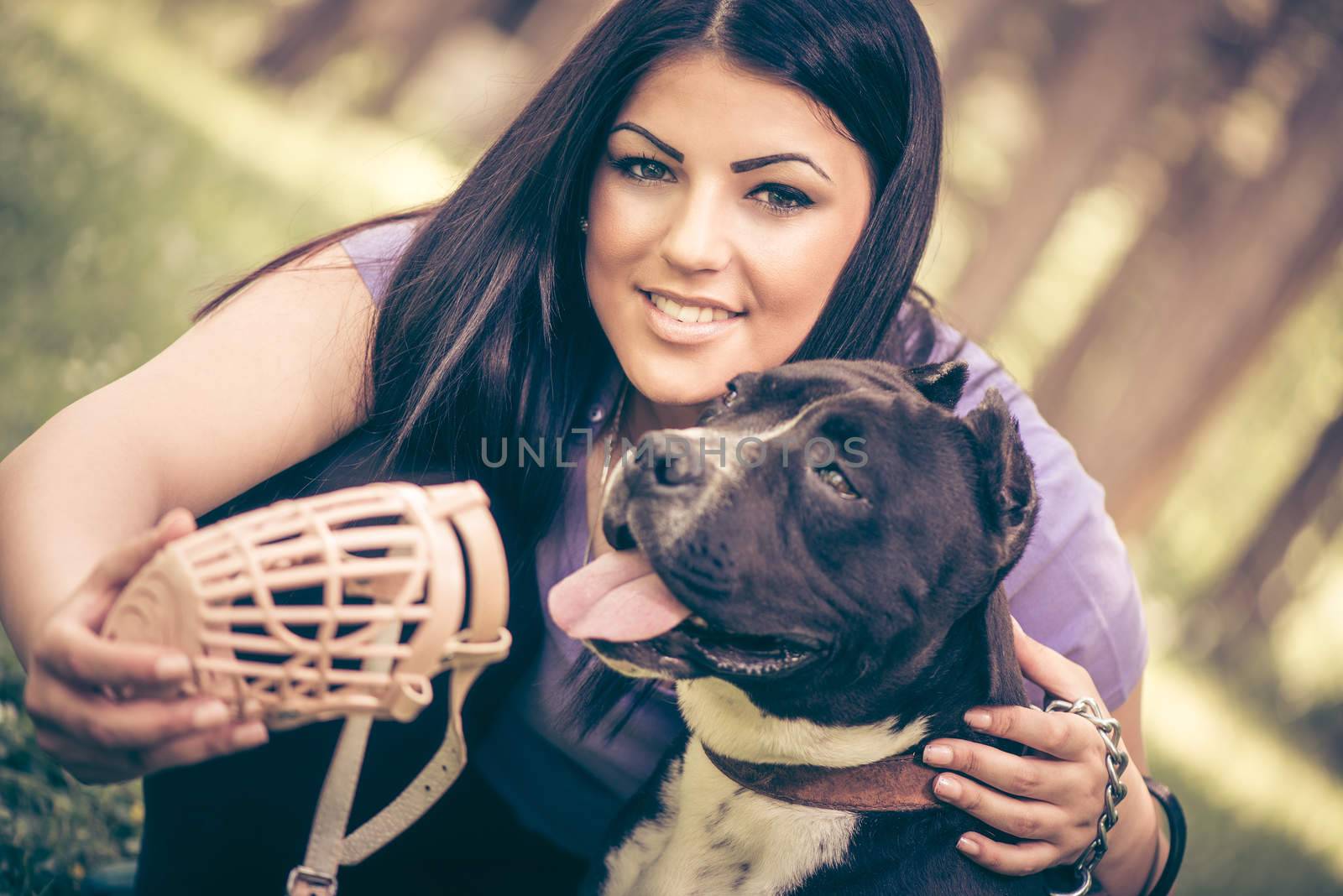 Beautiful young woman puts muzzle basket to her cute stafford terrier in the park .