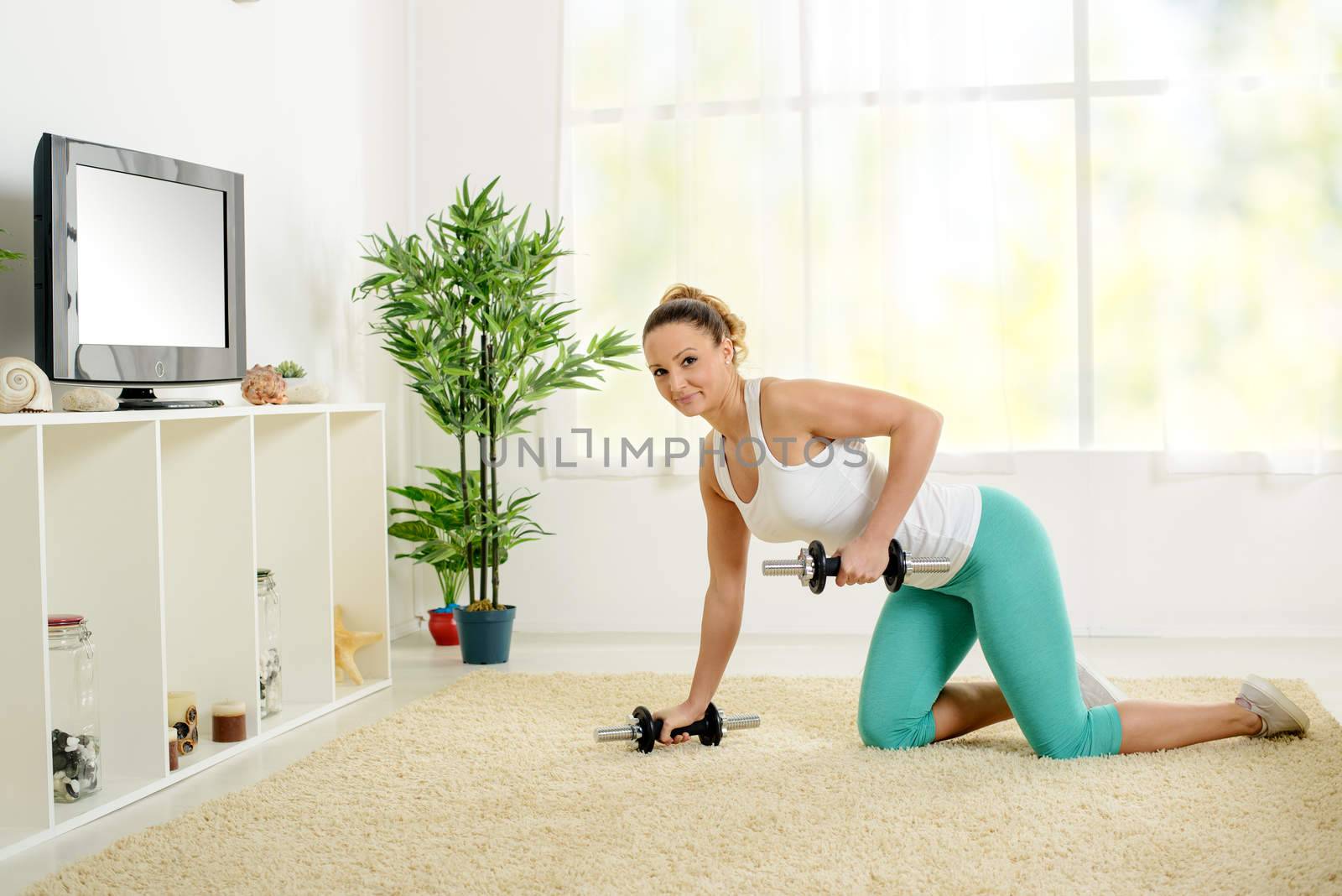 Beautiful young woman doing exercise to strengthen backs with dumbbells at home in front of the tv.