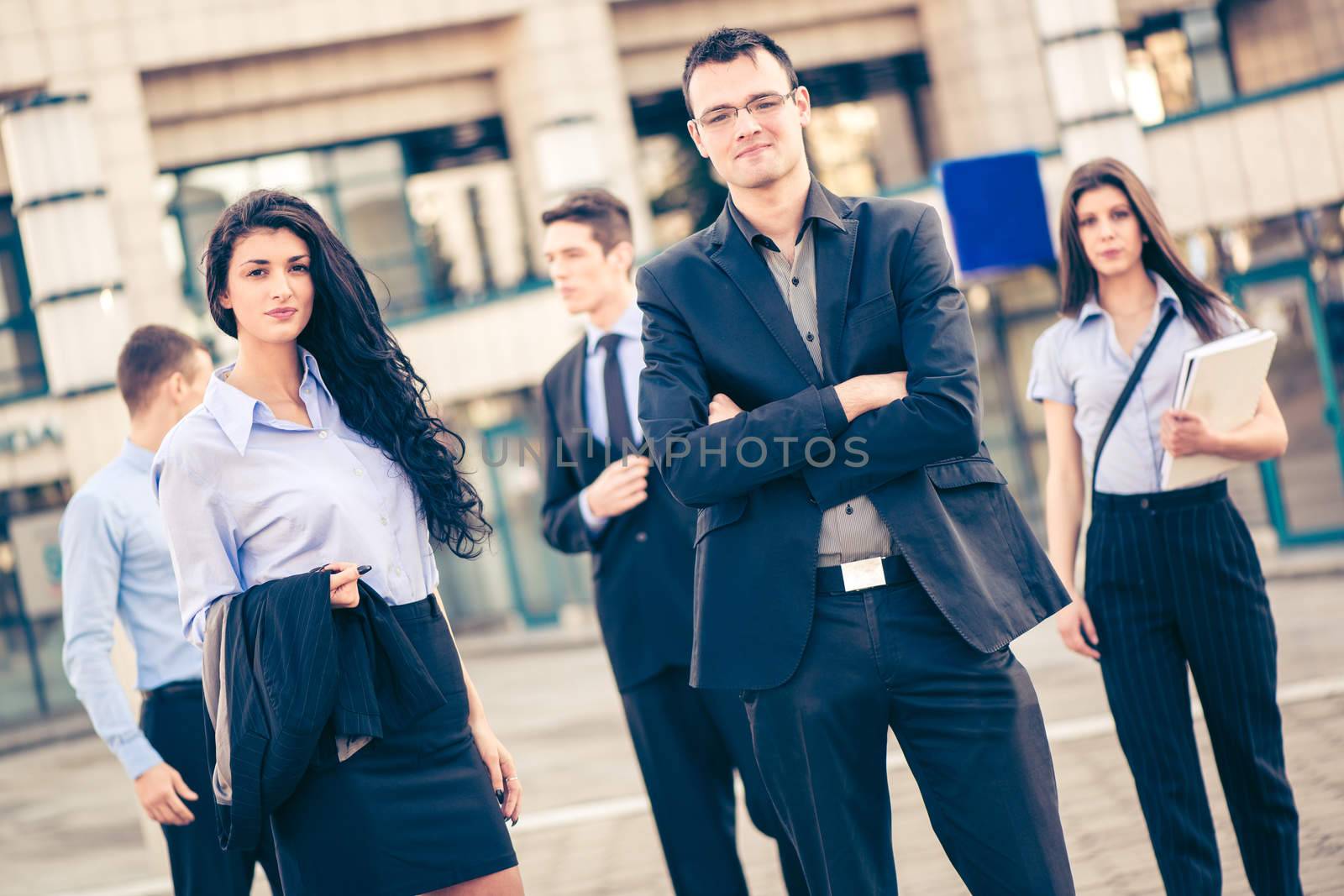 Young businessman and businesswoman with his team in the background standing in front of office building and looking at camera.