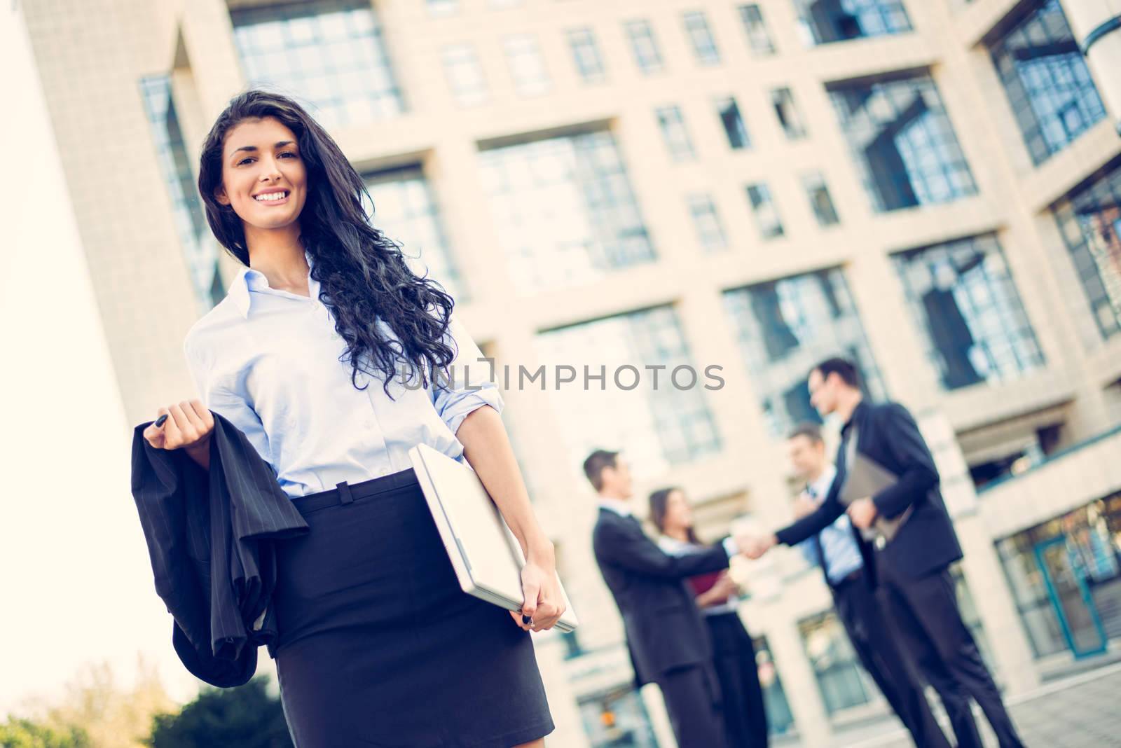 A young businesswoman standing in front of office building separated from the rest of the business team, holding laptop and looking at the camera.