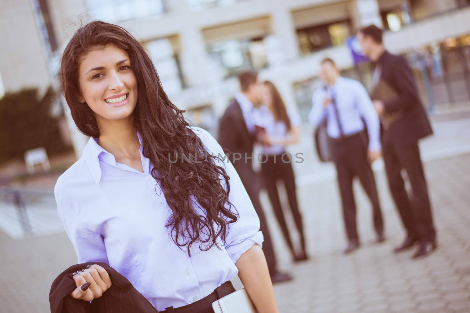 Portrait a young businesswoman standing in front of office building separated from the rest of the business team. Looking at the camera. 