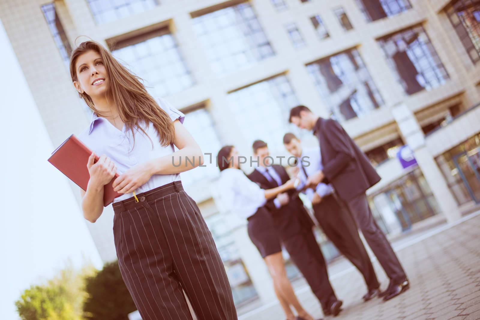 Young businesswoman with planner in her hands, standing in front of office building separated from the rest of the business team.