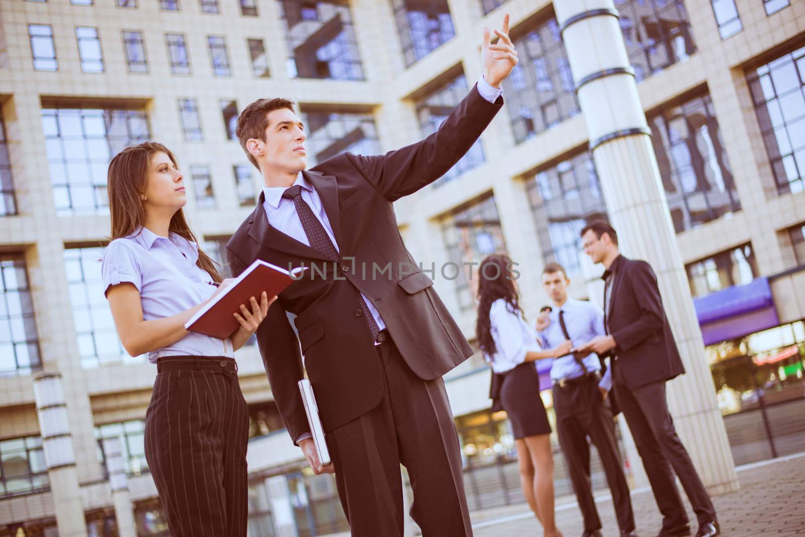 Young businessman and his secretary standing in front of office building.  Businessman hand pointing to the heights, and the secretary keeps the planner.