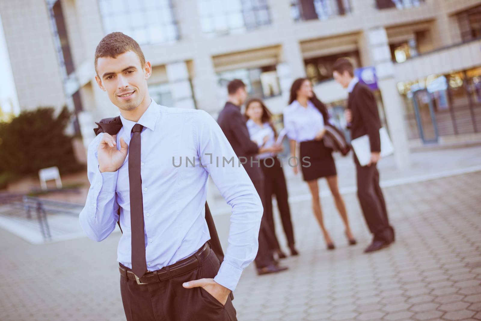 Young smiling businessman standing in front of office building separated from the rest of the business team. Looking at camera.