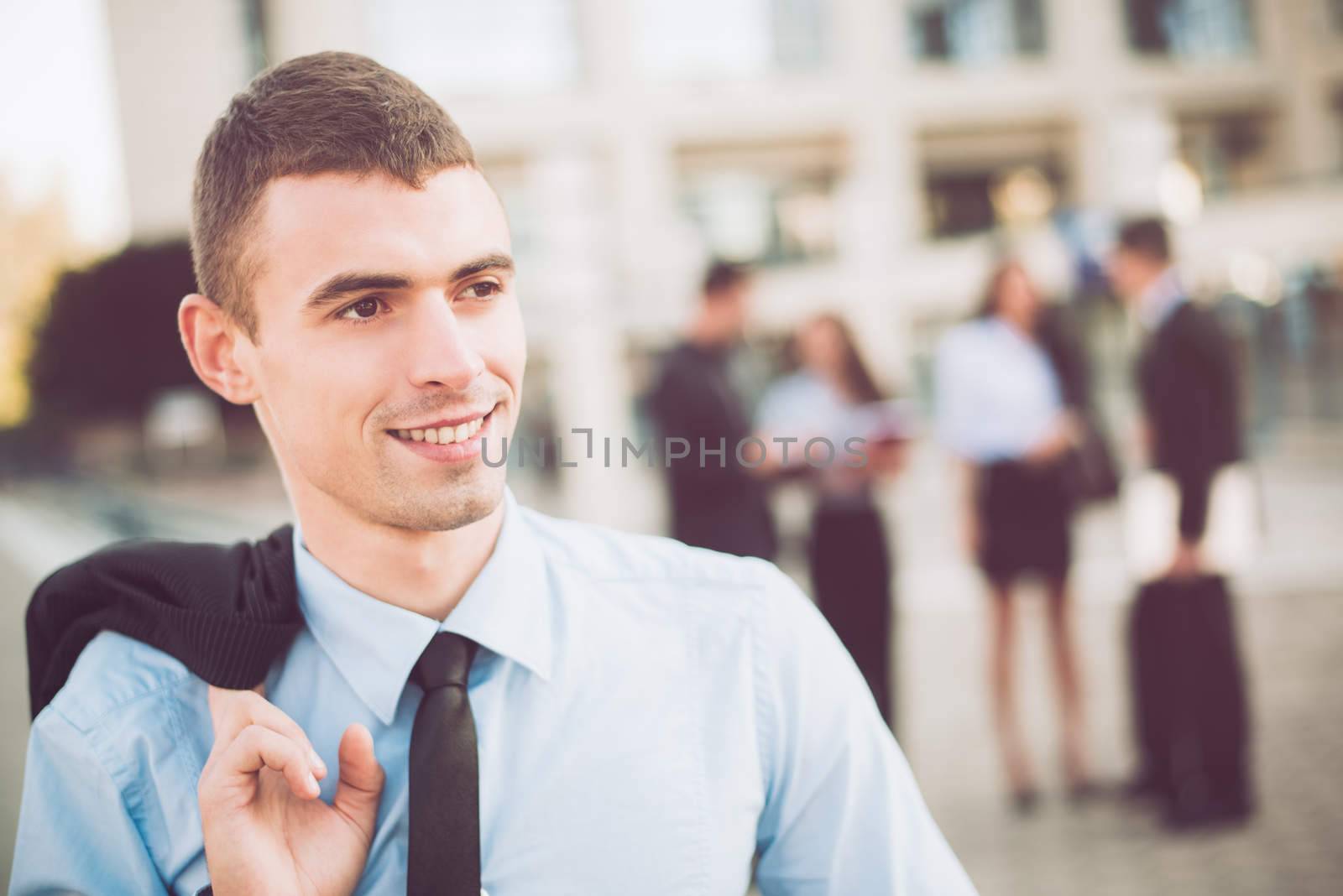 Portrait of a young smiling businessman standing in front of office building separated from the rest of the business team.