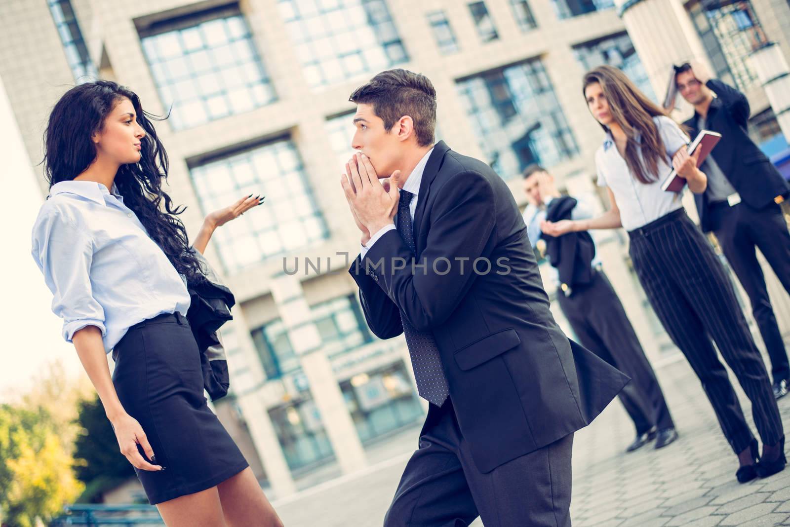 Young handsome, elegantly dressed businessman on the street in front of office building, kneeling with folded hands in front of a young attractive business woman while they watching colleagues.