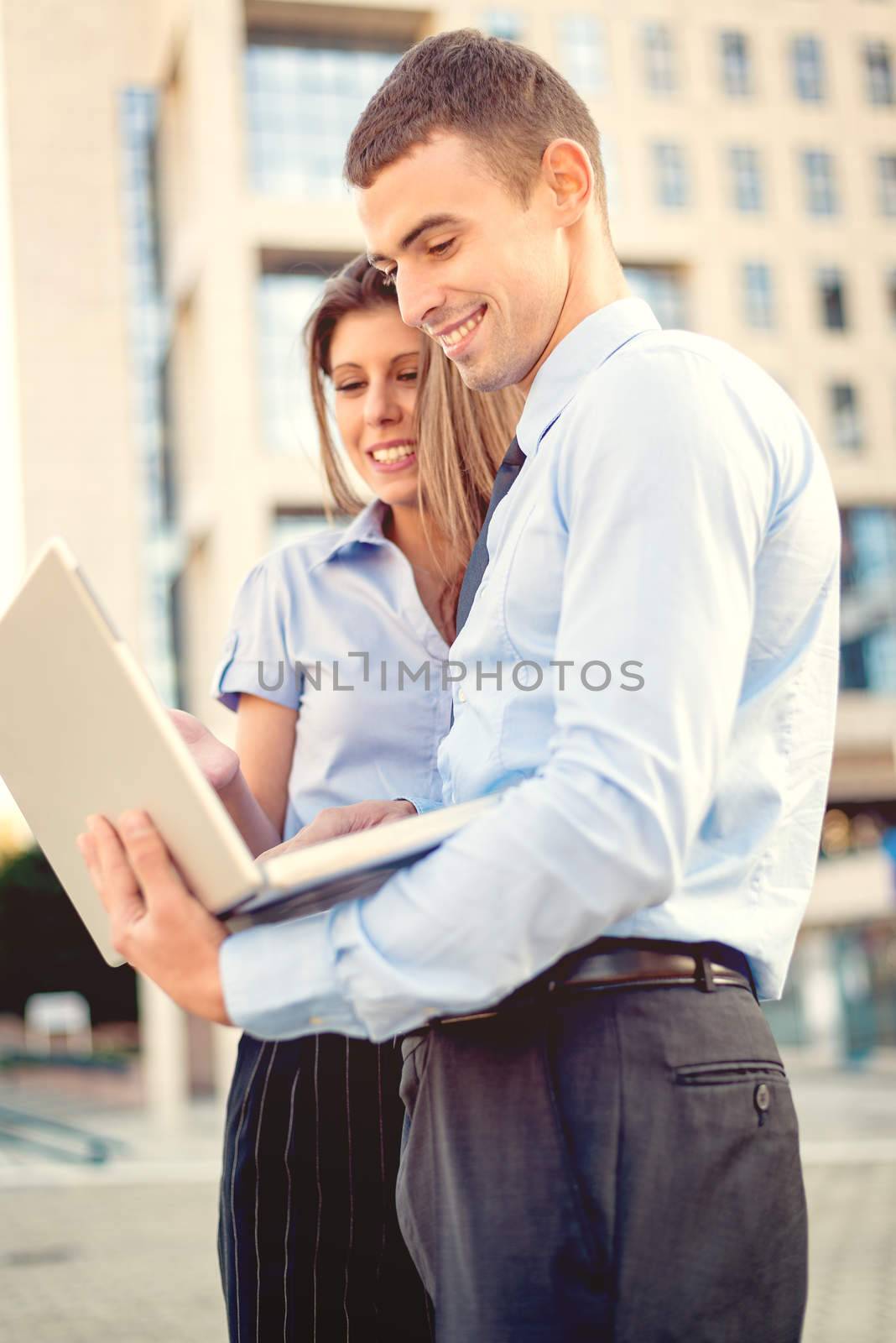 Smiling young businessman and businesswoman in front of office building looking at the laptop, while from behind the sun shines.