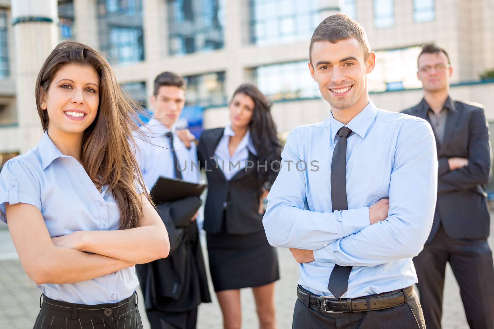 A young and pretty businesswoman and businessman with their team in the background standing in front of the company by looking at the camera and smiling.