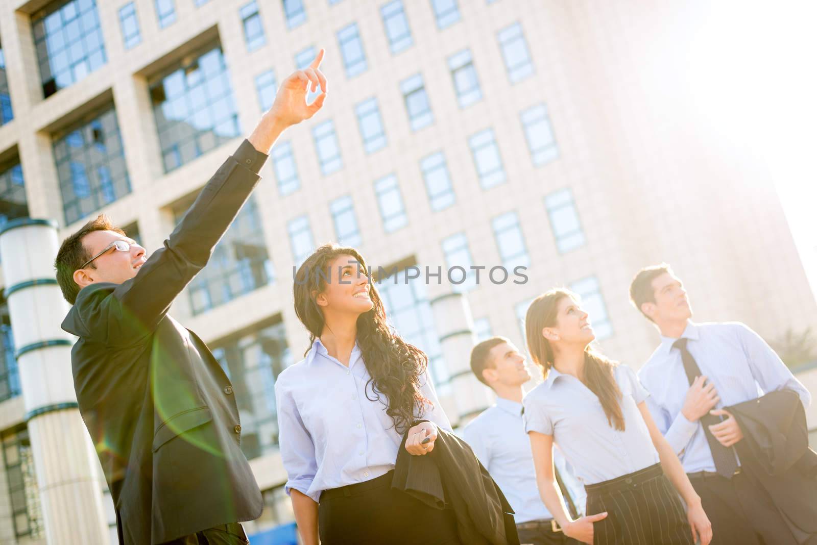 The young businessman in front of office building outstretched fingers of the hand, aimed at heights, shows his young business team motivating them to new business success, while the sun shines from behind.