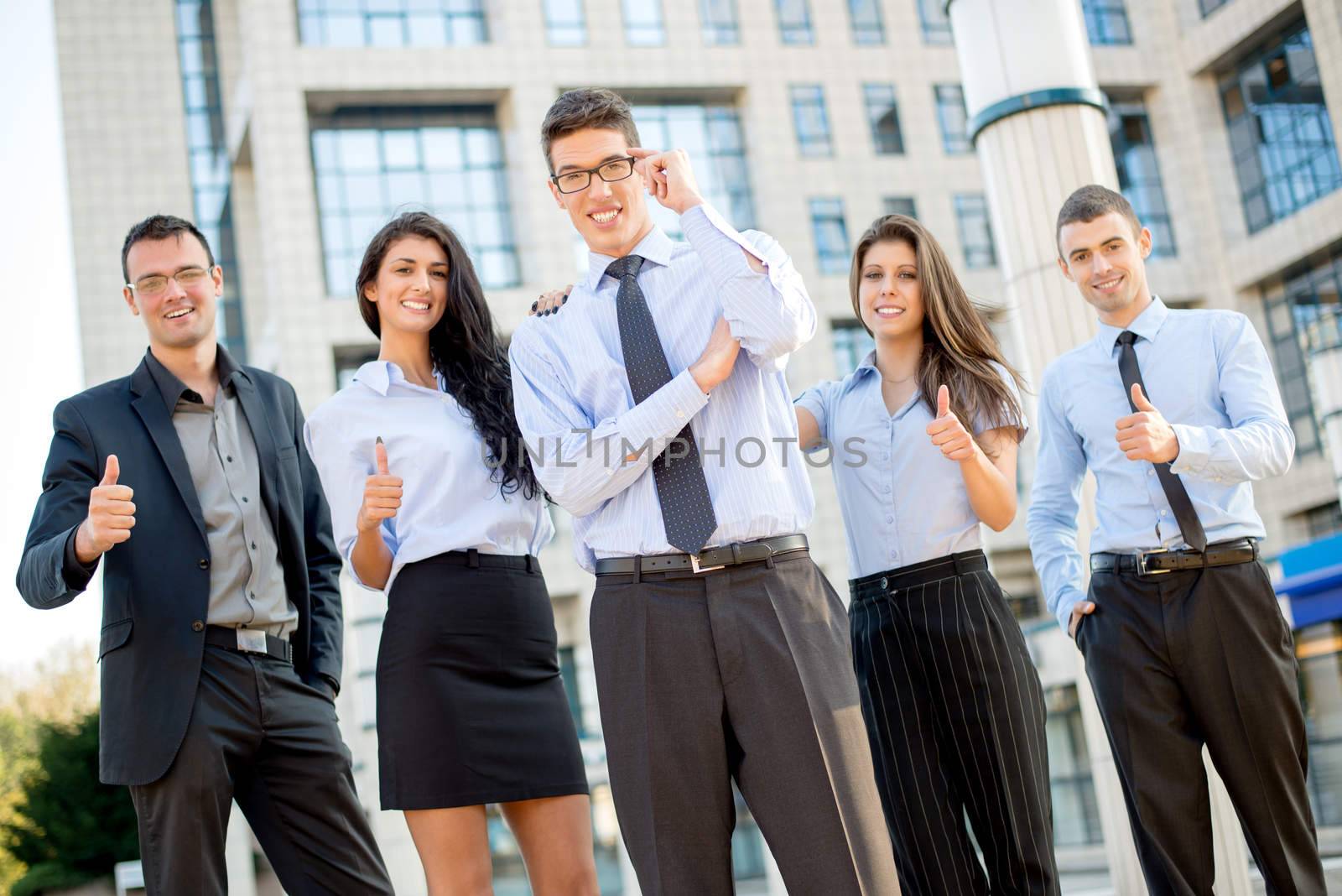 Young businessman with glasses, elegantly dressed standing outside in front of office building while behind him stands his young business team with thumbs up.