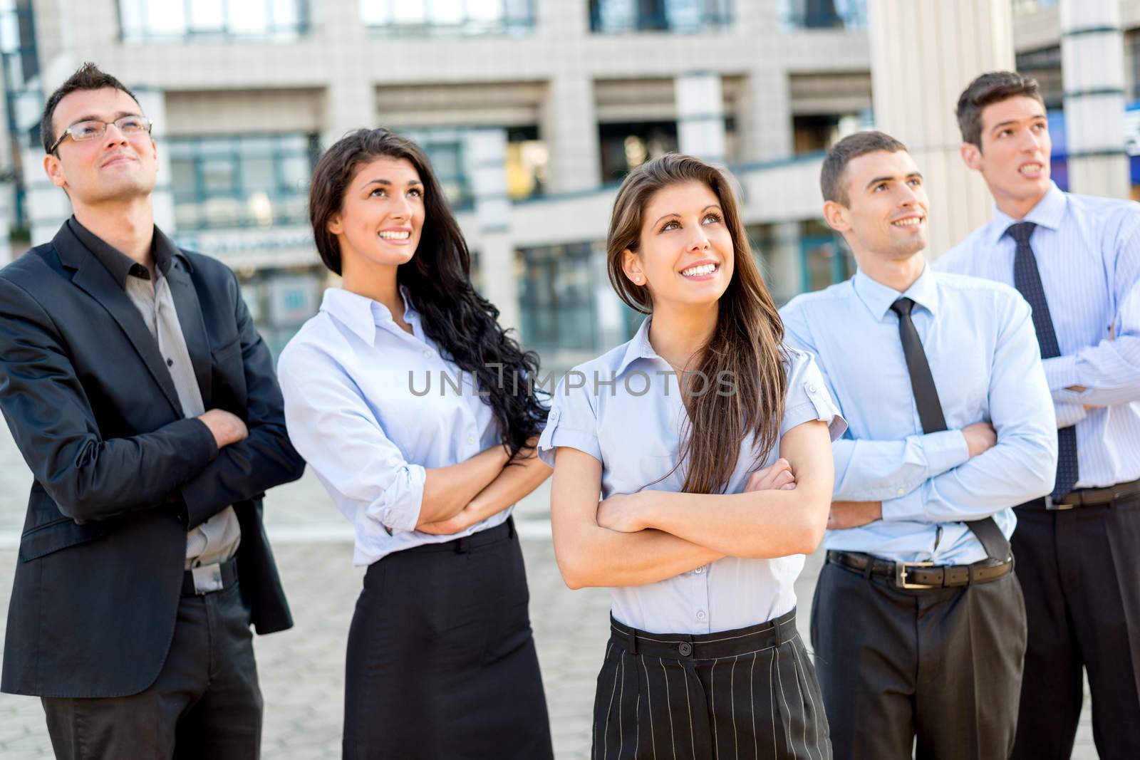 A small group of young business people standing in front of office building with arms crossed and with a smile on their faces looking in height.