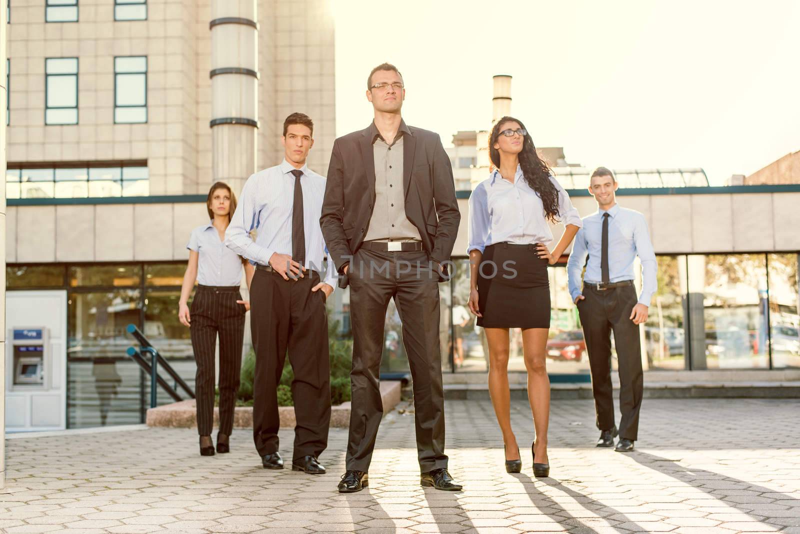 Young businessman, elegantly dressed with his hands in his pockets, standing proudly with his team of young businesswomen and businessmen in front of office building illuminated backlit sun.