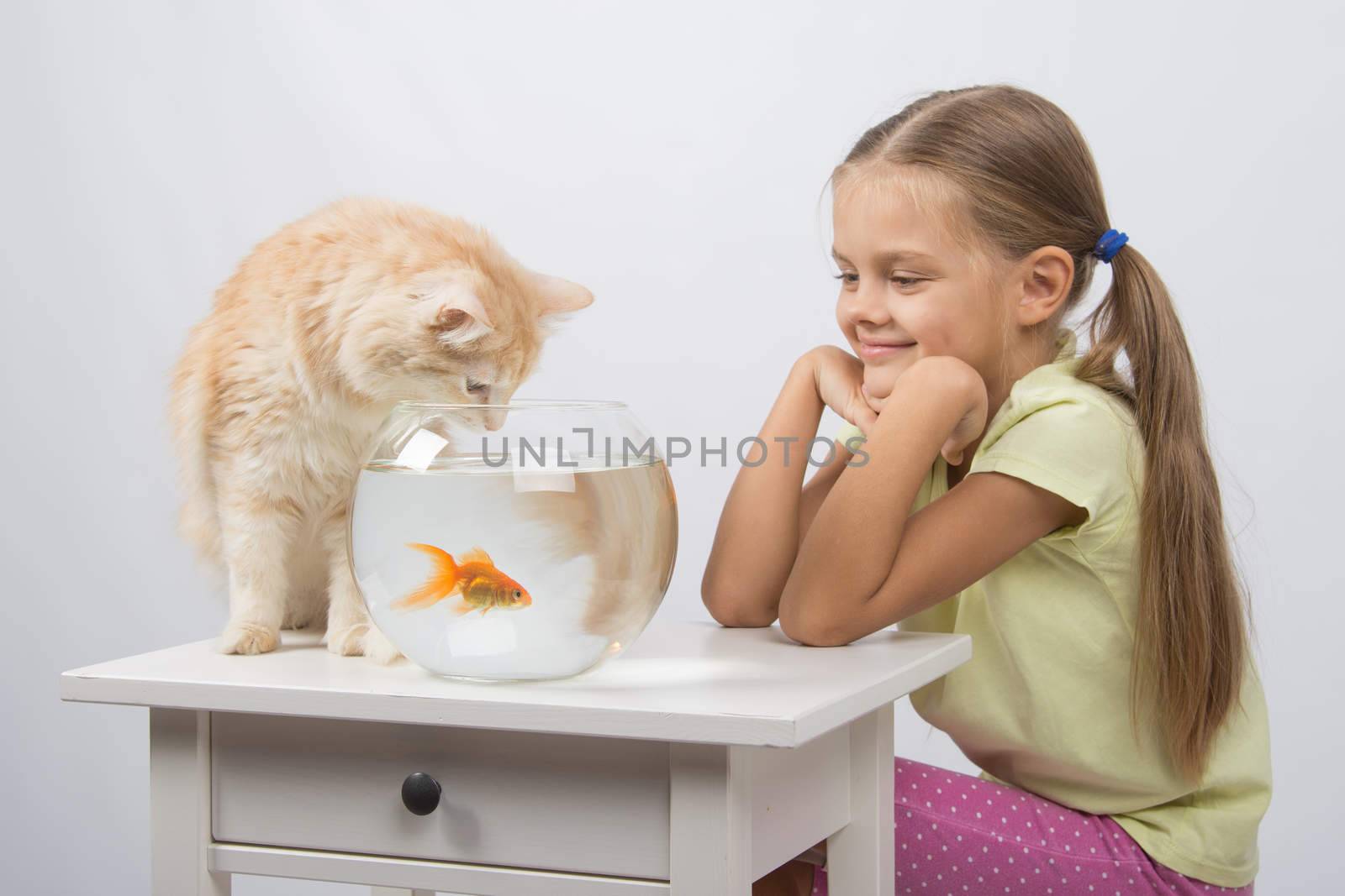 Happy six year old girl and a cat sitting at the table at the fishbowl with goldfish