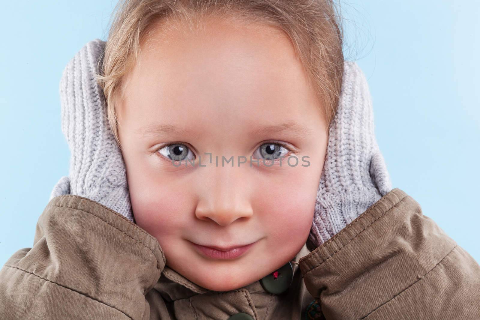 Little cute girl covering her ears  with hands in gloves against light blue background. Winter season.