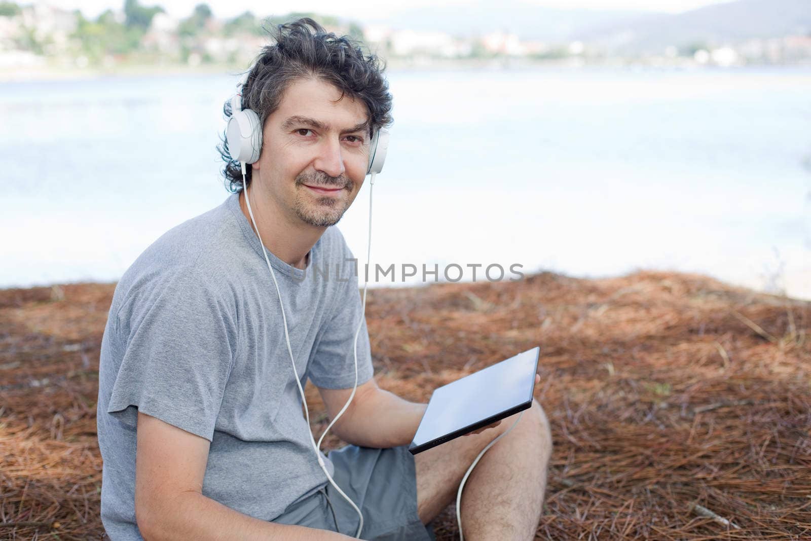 young man holding a tablet with headphones, at the beach